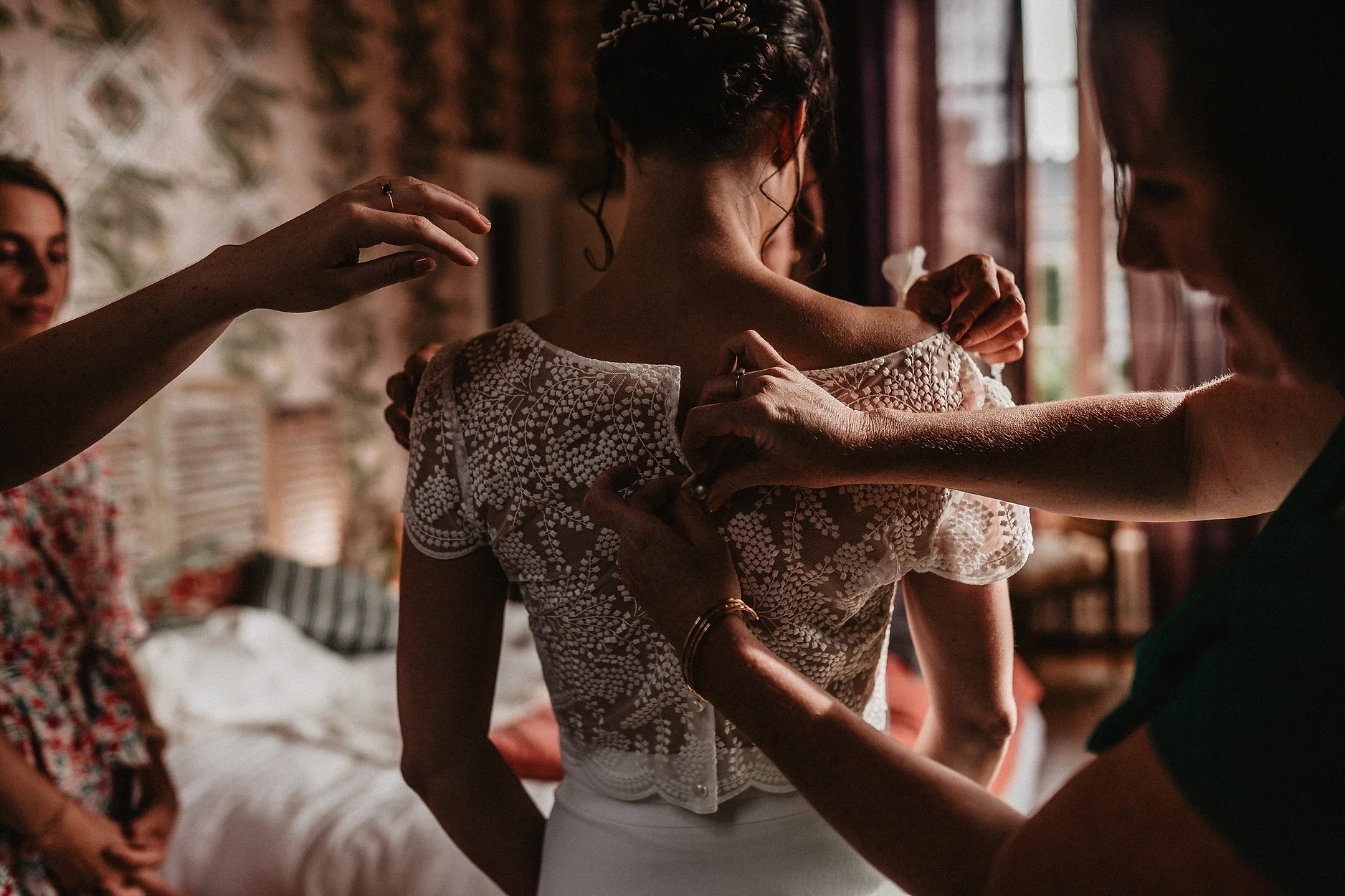 Groupe de femmes aidant une femme à enfiler une robe de mariée en dentelle dans une pièce lumineuse.