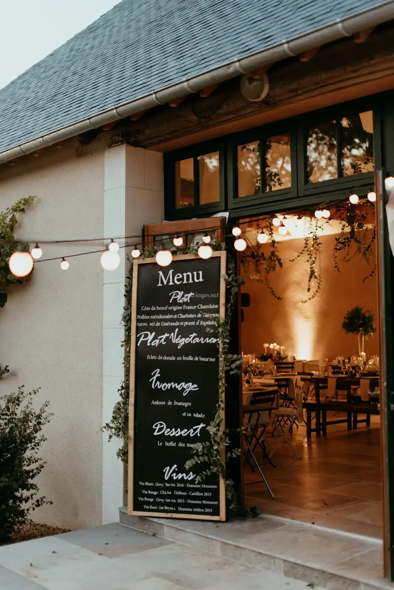 Panneau de menu d'un restaurant français avec des guirlandes lumineuses extérieures, et une salle à manger chaleureuse à l'intérieur visible par la porte ouverte.