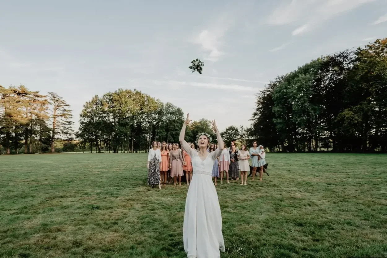 Une jeune femme en robe blanche lance un bouquet de fleurs en l'air devant un groupe de femmes dans un parc.