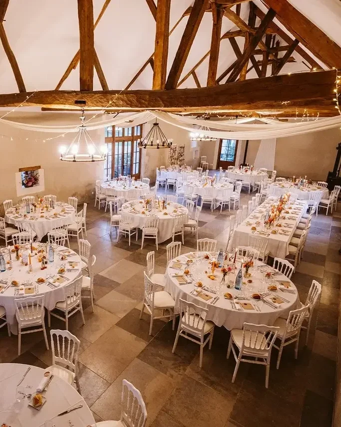 Salle de réception décorée pour un événement avec des tables rondes et rectangulaires avec nappes blanches, vaisselle, couverts, verre, et éléments de décoration au centre des tables.