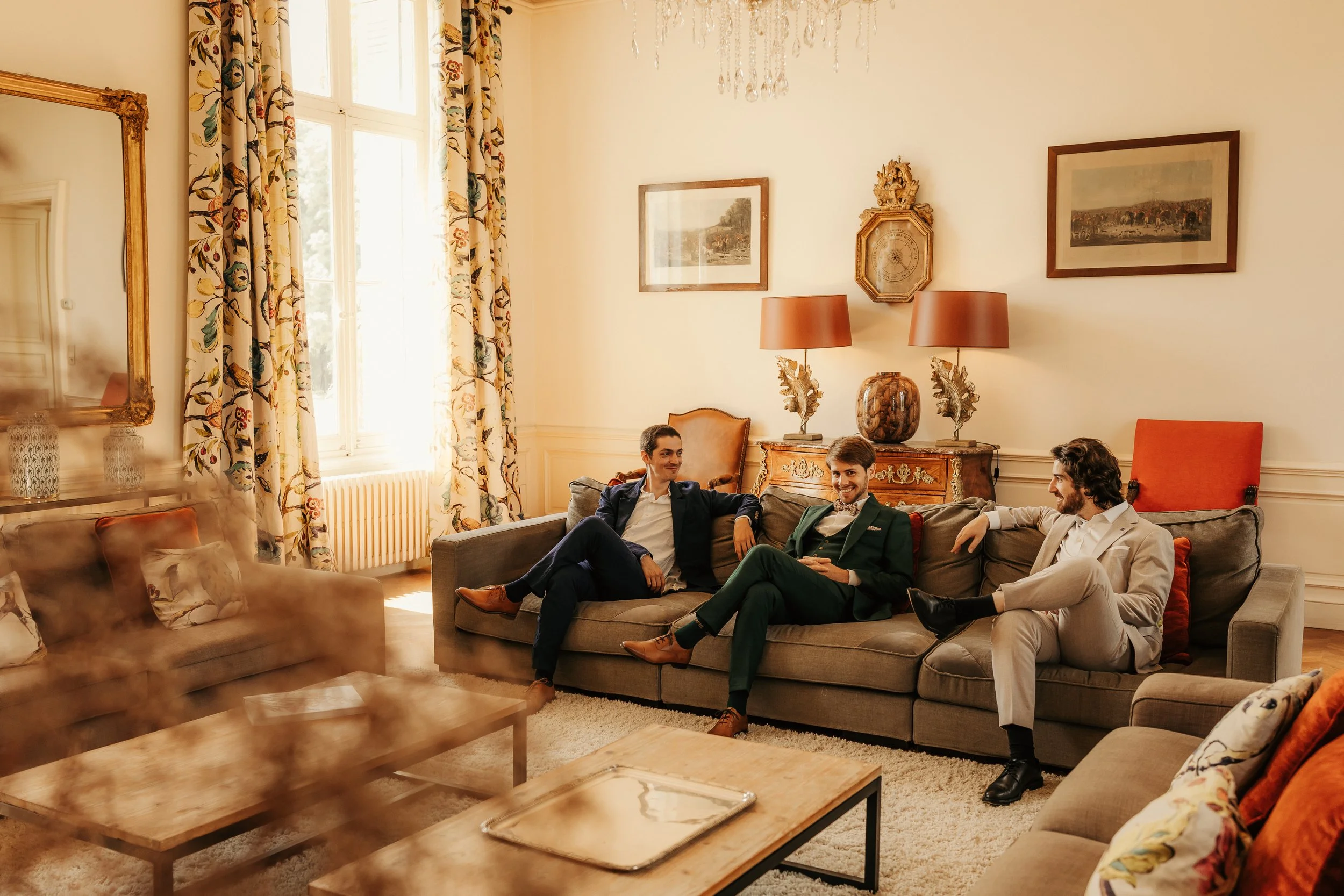 Trois hommes assis sur un canapé dans un salon élégant, discutant et souriant, avec des décorations classiques et des lampes en arrière-plan.