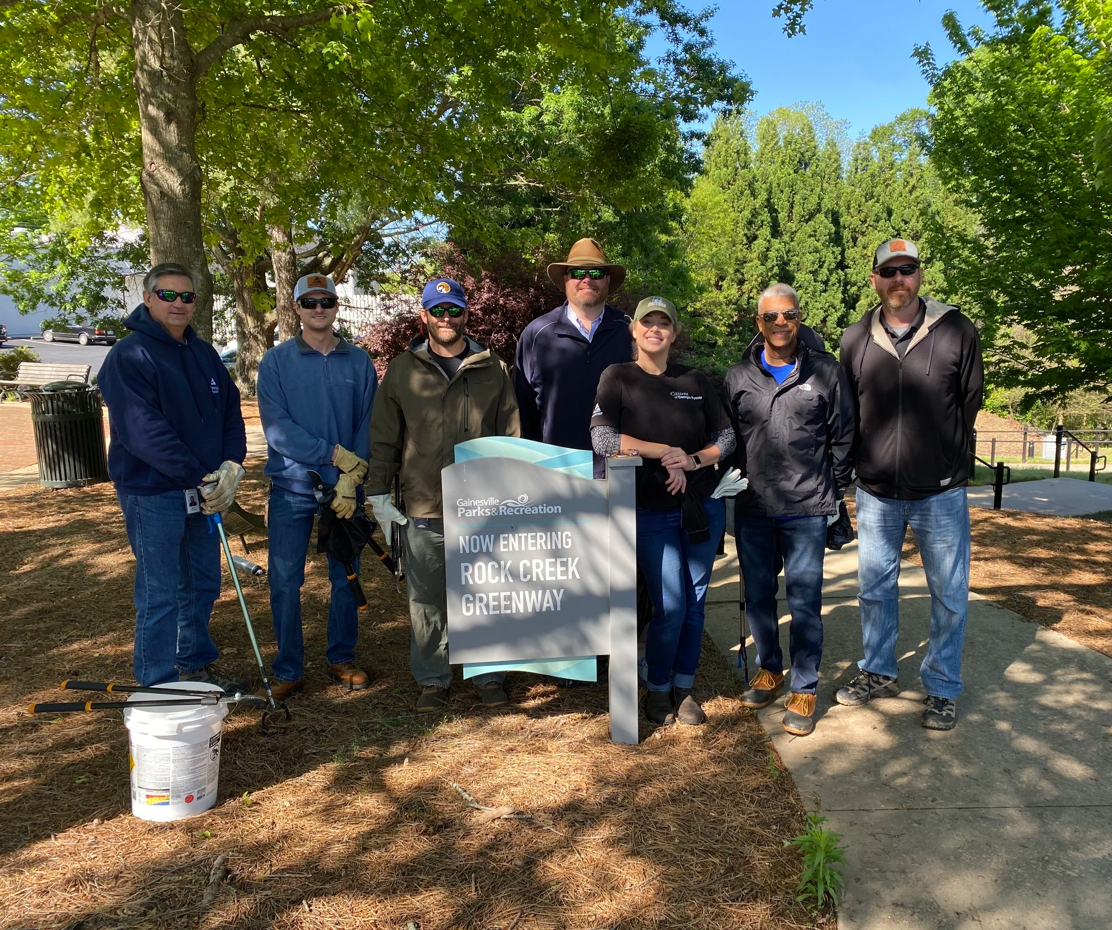 Group of seven people standing outdoors near a park sign that reads 'Now Entering Rock Creek Greenway,' with trees in the background on a sunny day.