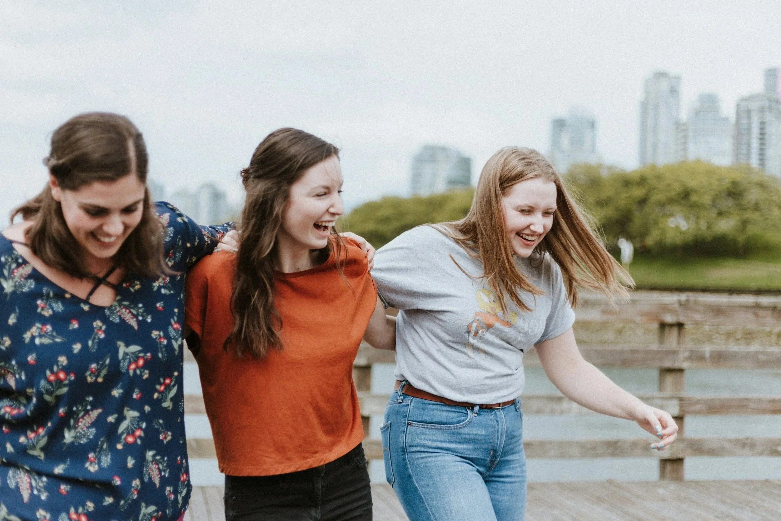 Three young women walking together outdoors on a wooden path, smiling and laughing.
