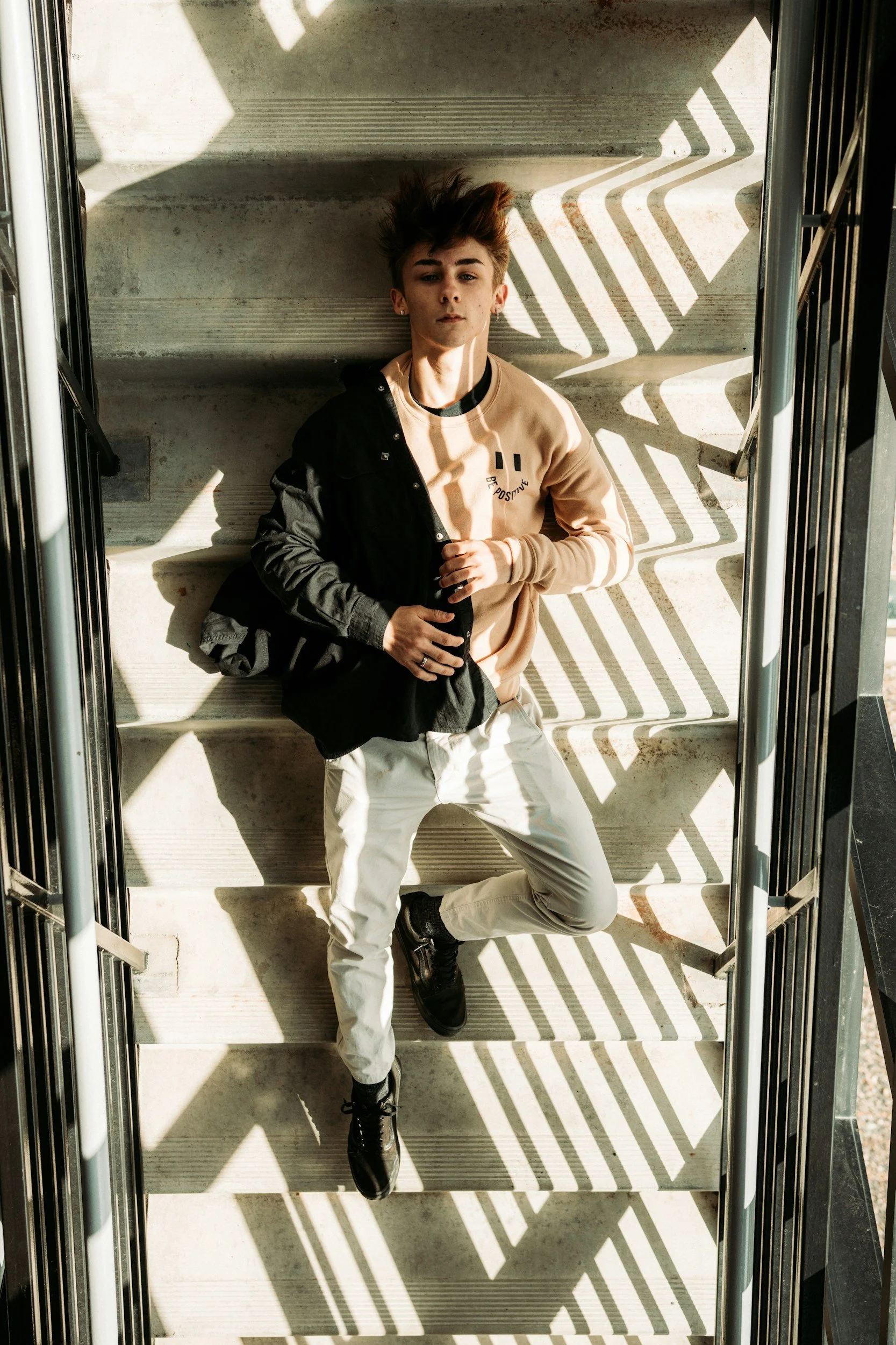 A young person with styled hair lying on stairs, wearing a beige and black jacket and white pants, with shadows cast by railing patterns.