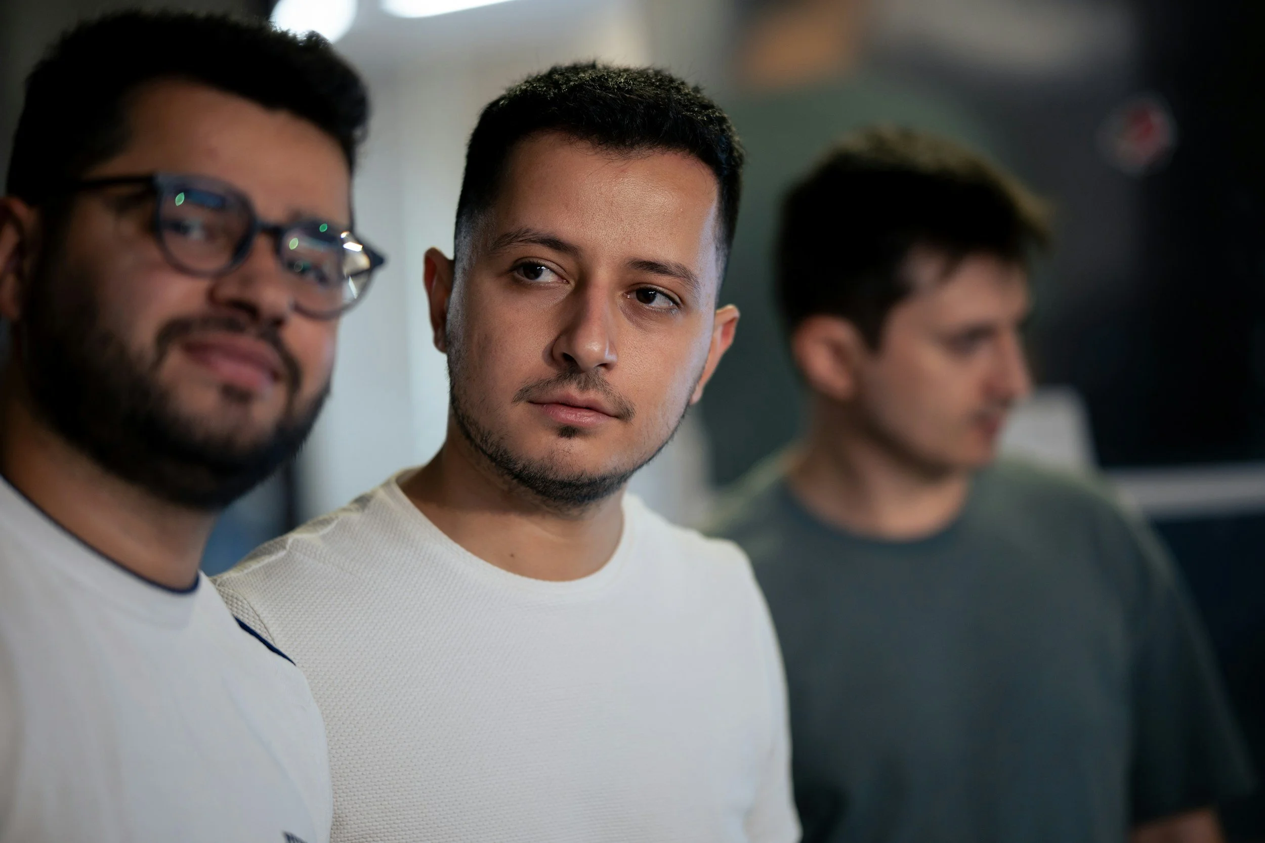 Three young men standing in a line, with the man in the middle in focus, looking slightly to the side, in an indoor setting.
