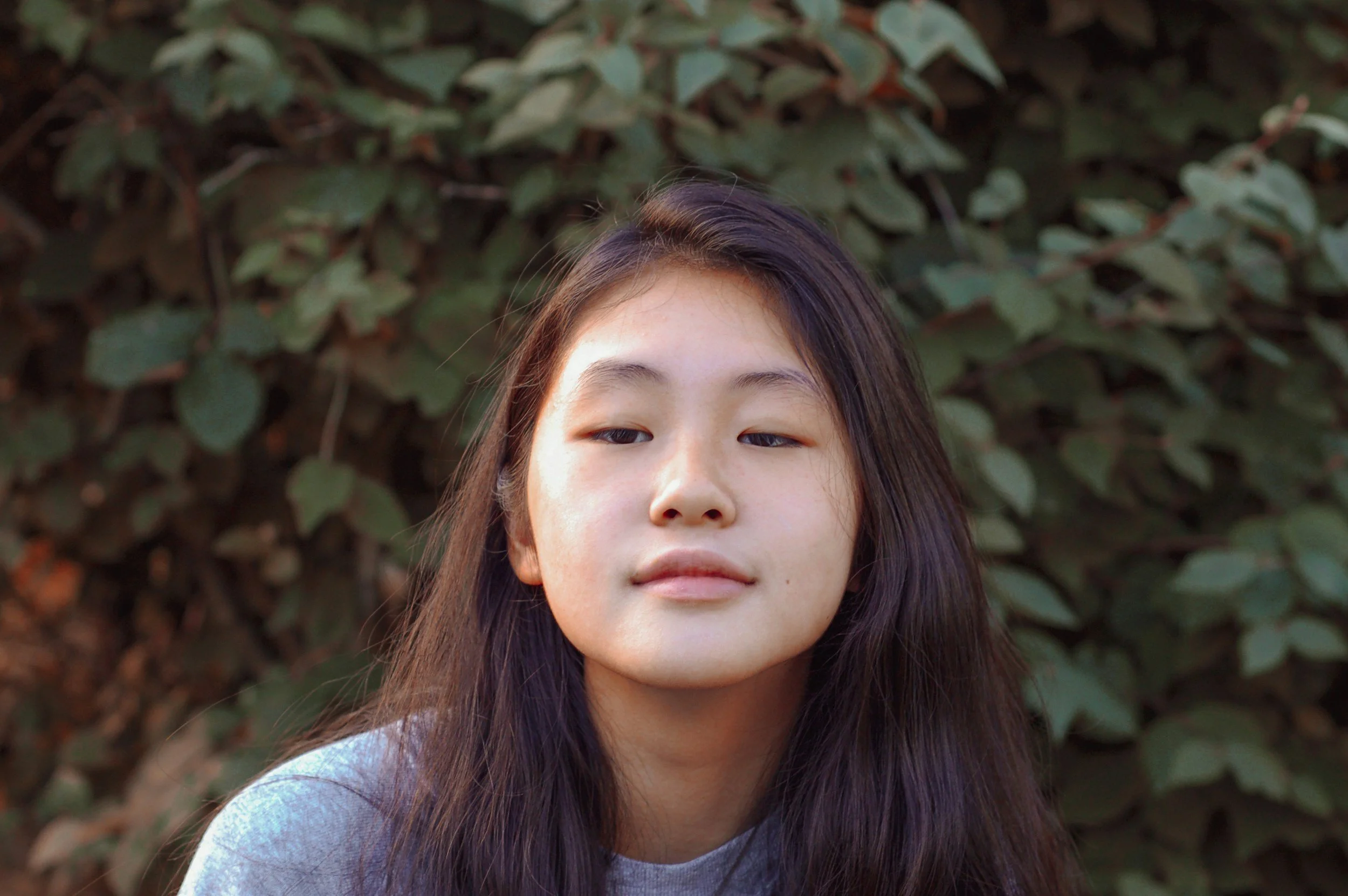 A young girl with long dark hair, wearing a gray shirt, standing outdoors in front of dense green foliage.