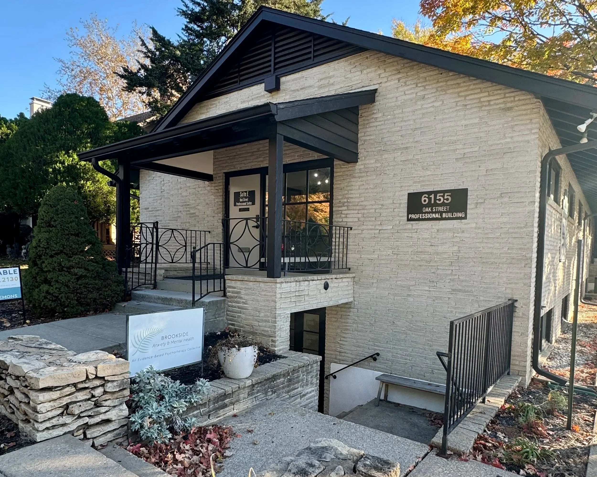 Outside view of a beige brick professional building with black accents at 6155 Oak Street. A ramp and stairs lead to the entrance with a black railing. A sign reads 'Brookside Anxiety & Mental Health' near plants and a small garden area. Fall foliage
