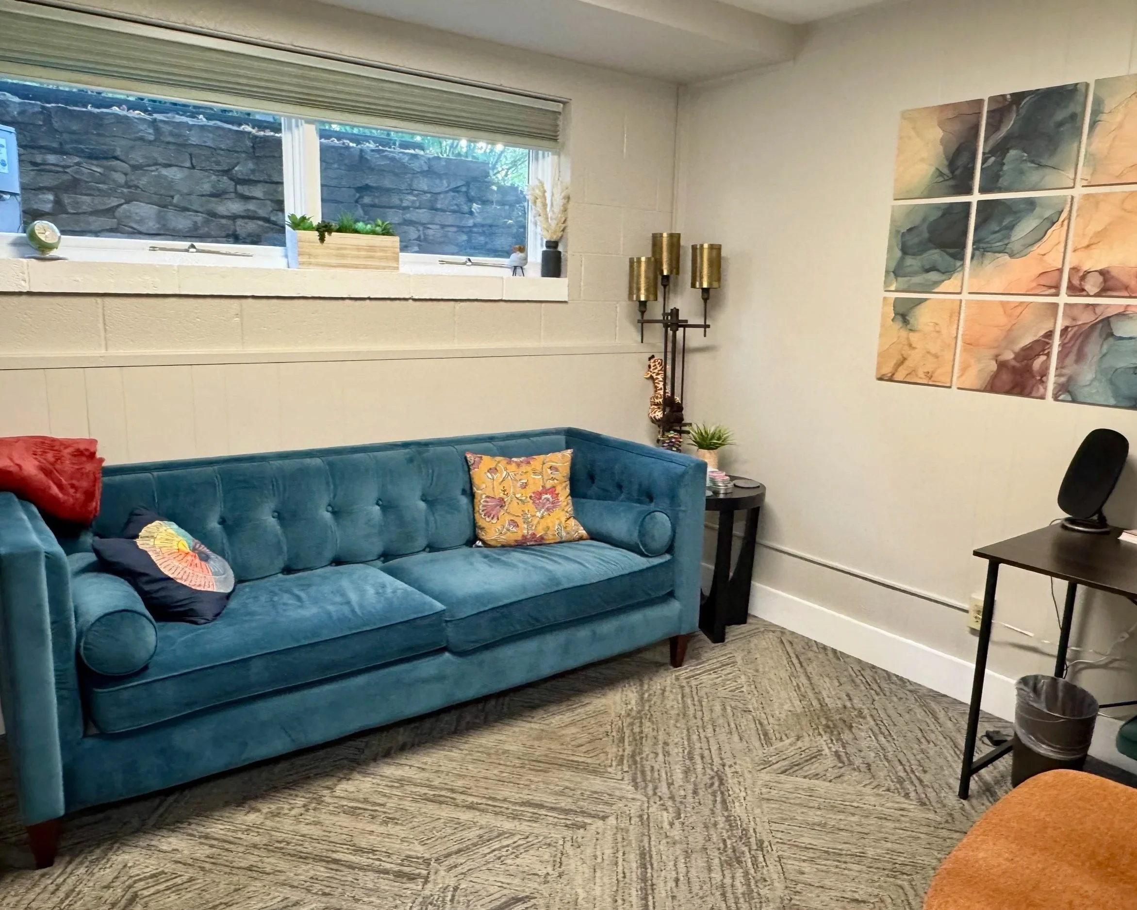 Living room with a teal velvet sofa, colorful decorative pillows, a side table with plants and a lamp, abstract wall art, and a window with a stone wall outside.