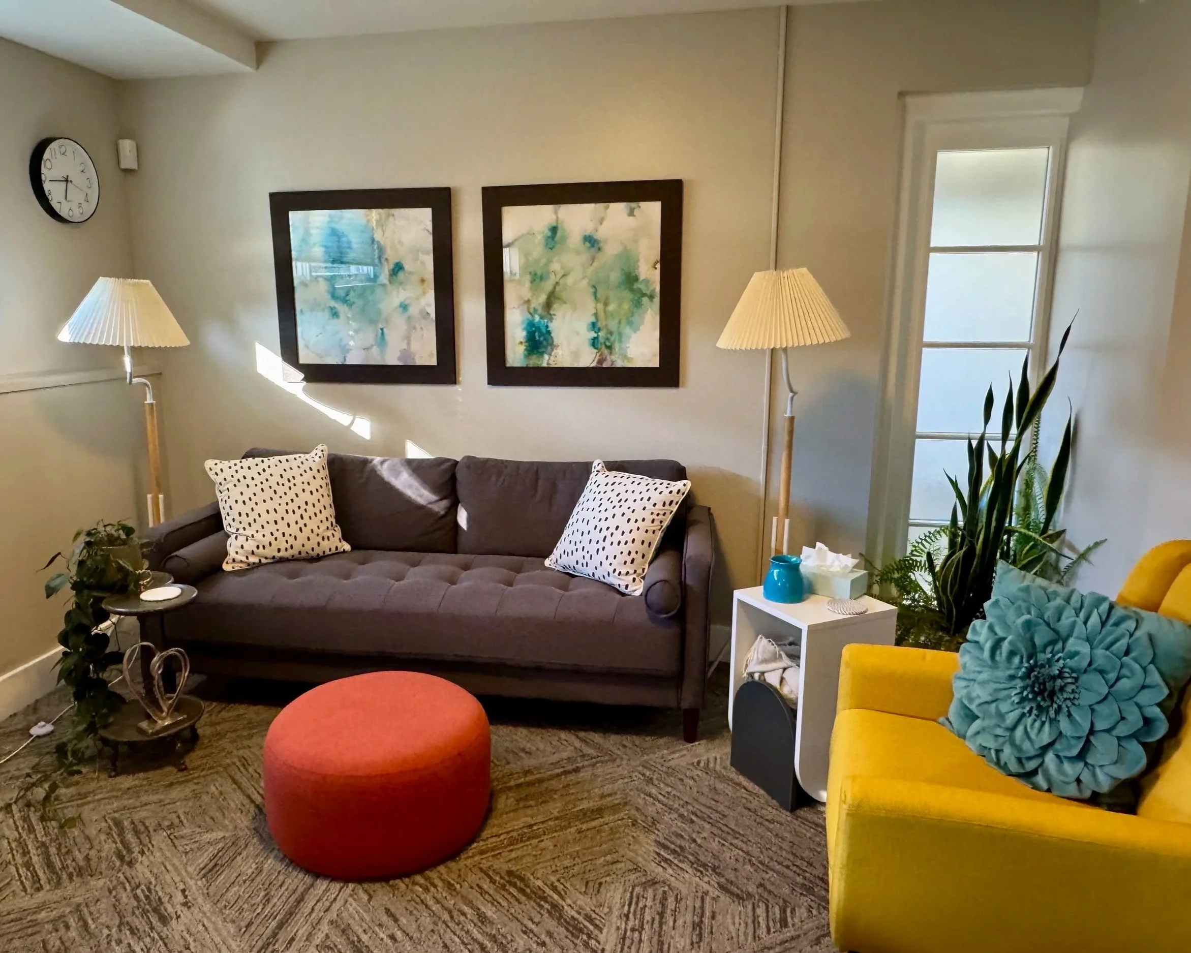 Cozy living room with gray sofa, yellow armchair, colorful accent pillows, abstract artwork, floor lamps, indoor plants, side table, and a textured carpet.