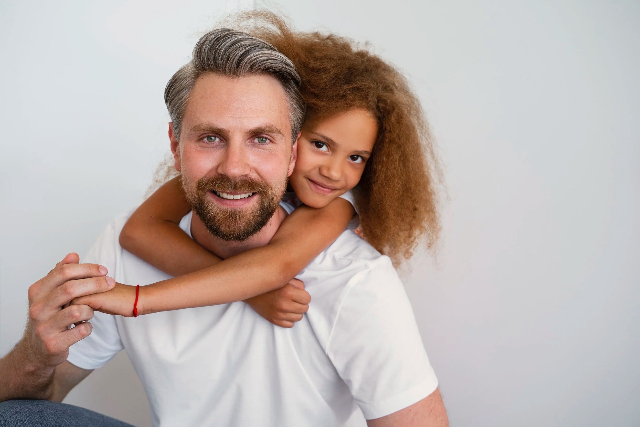 A man with a beard and a young girl with curly hair smiling, with the girl hugging the man's shoulders.