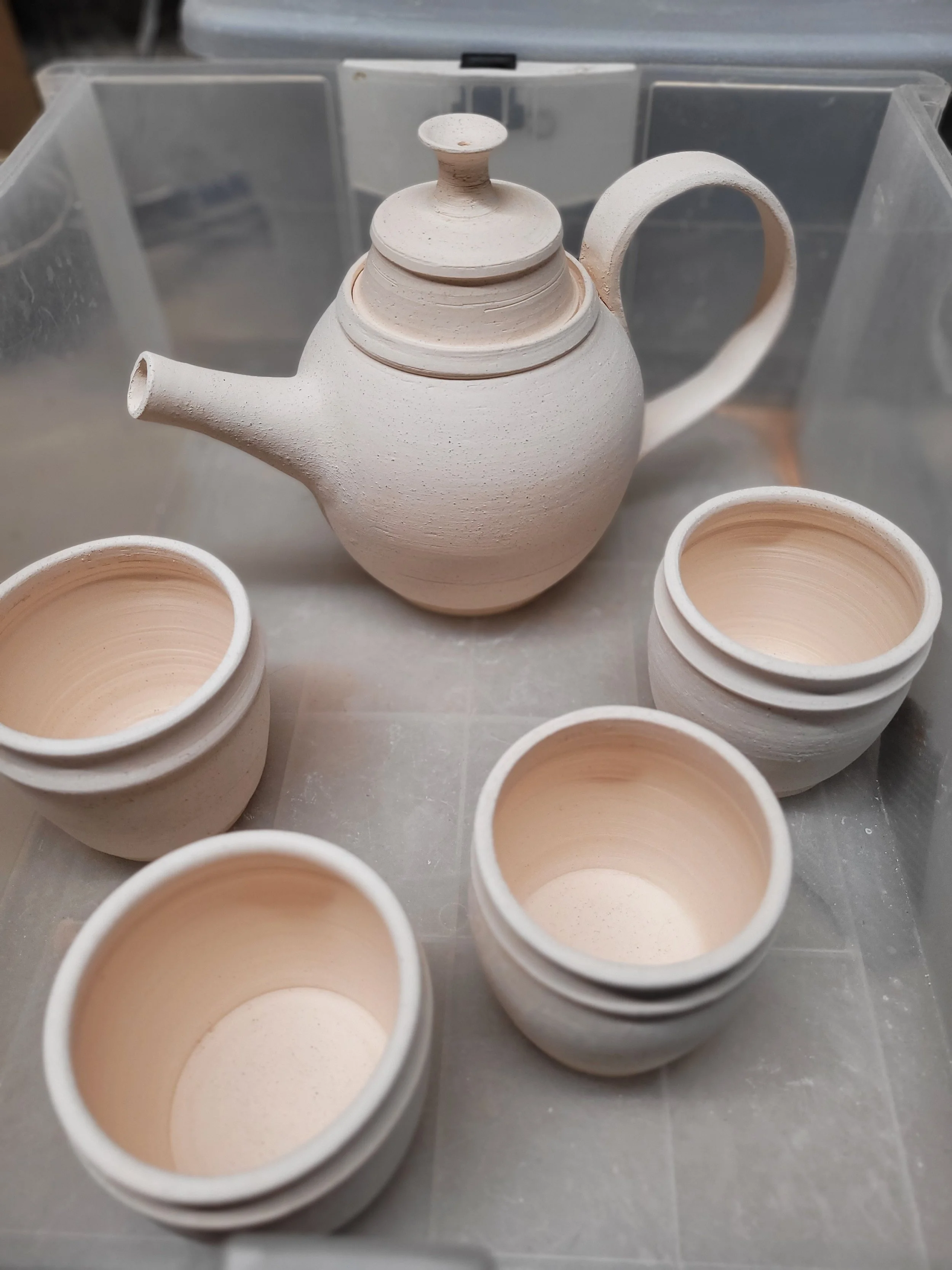 Unfinished white ceramic teapot with a lid, surrounded by five unglazed ceramic cups, all placed on a metallic surface.
