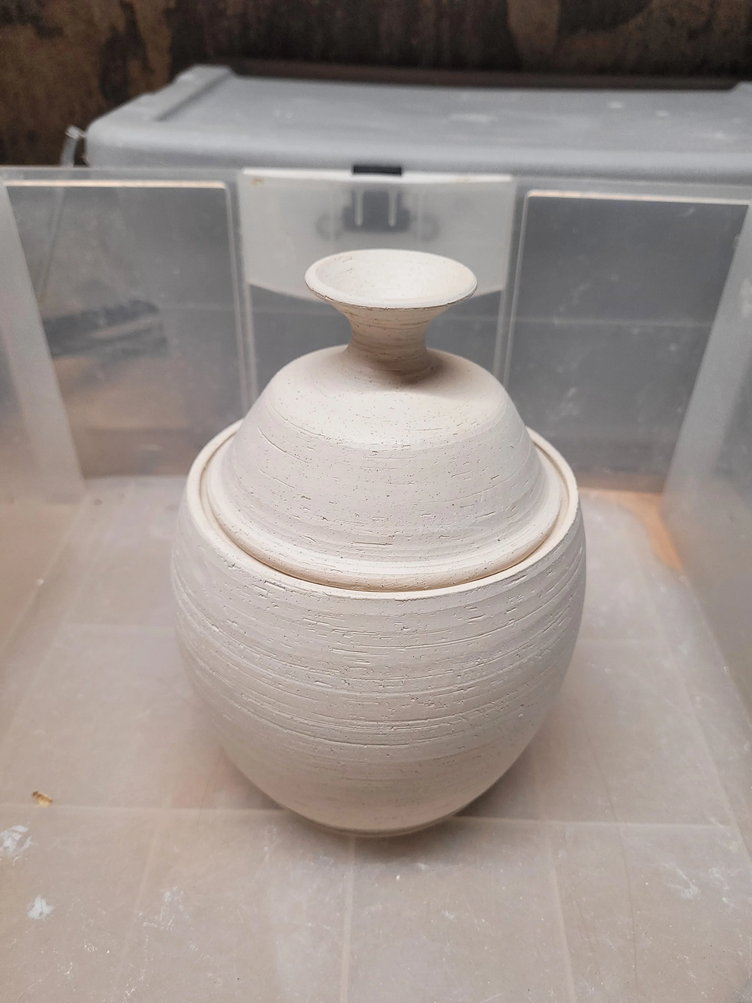 A piece of pottery in the shape of a rounded vessel with a short, wide neck and small, flat opening, placed inside a plastic box, with a blurred background of a brick wall.