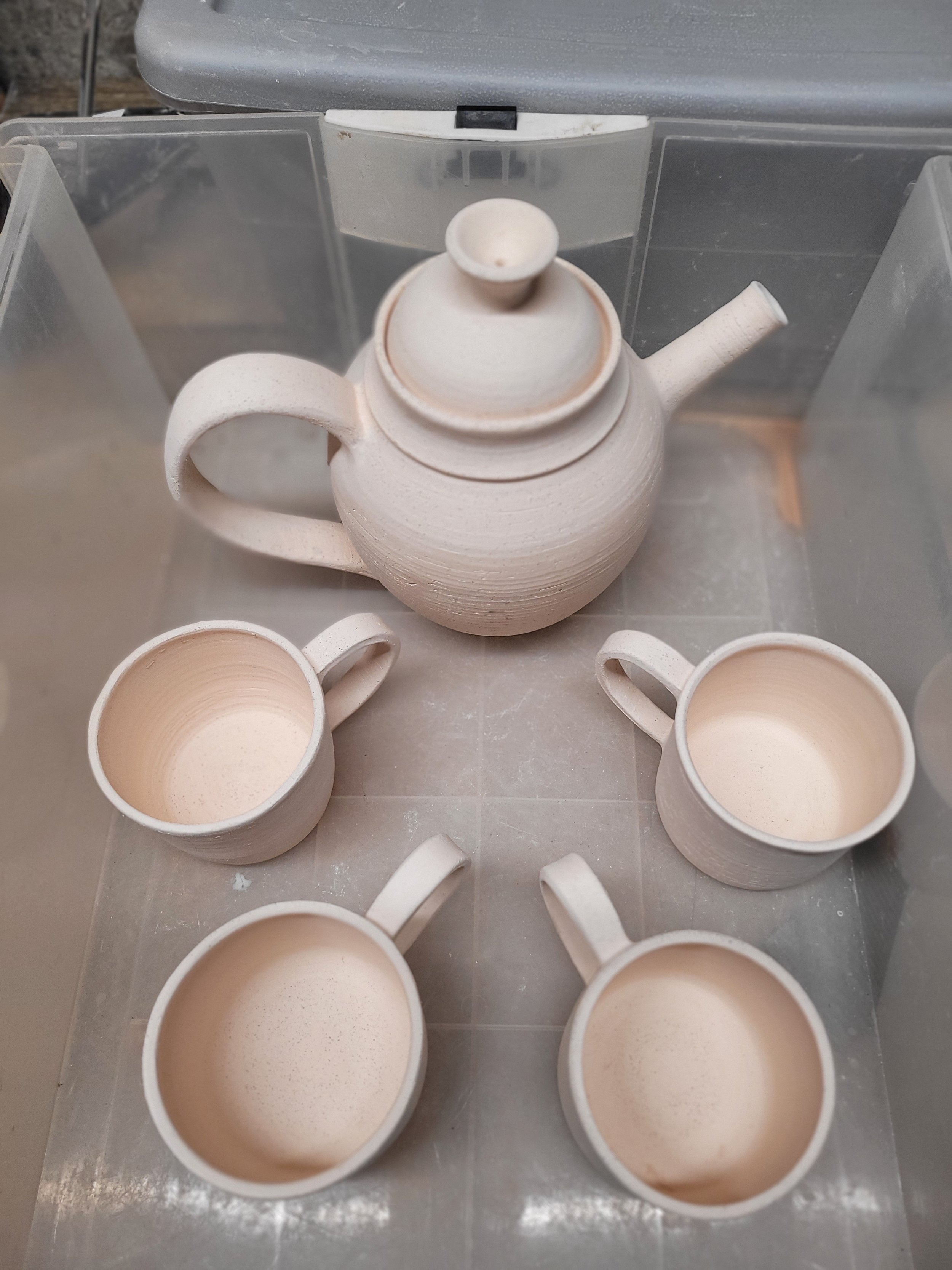 A ceramic teapot with a handle and spout, viewed from above inside a plastic storage bin, surrounded by several empty ceramic cups with handles.