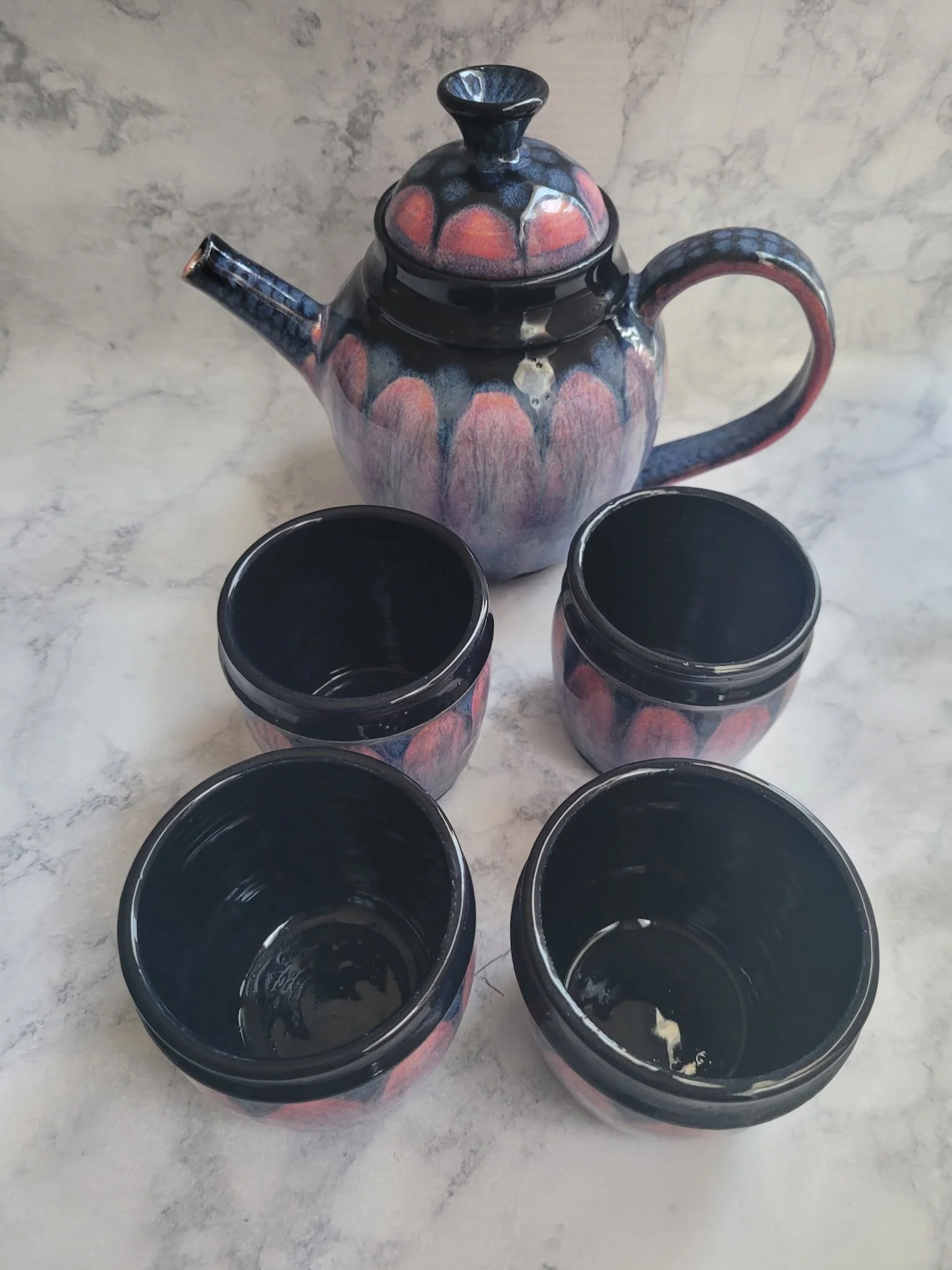 A ceramic teapot with a floral design and several matching cups, all with a dark interior and colorful exterior, placed on a white marble surface.