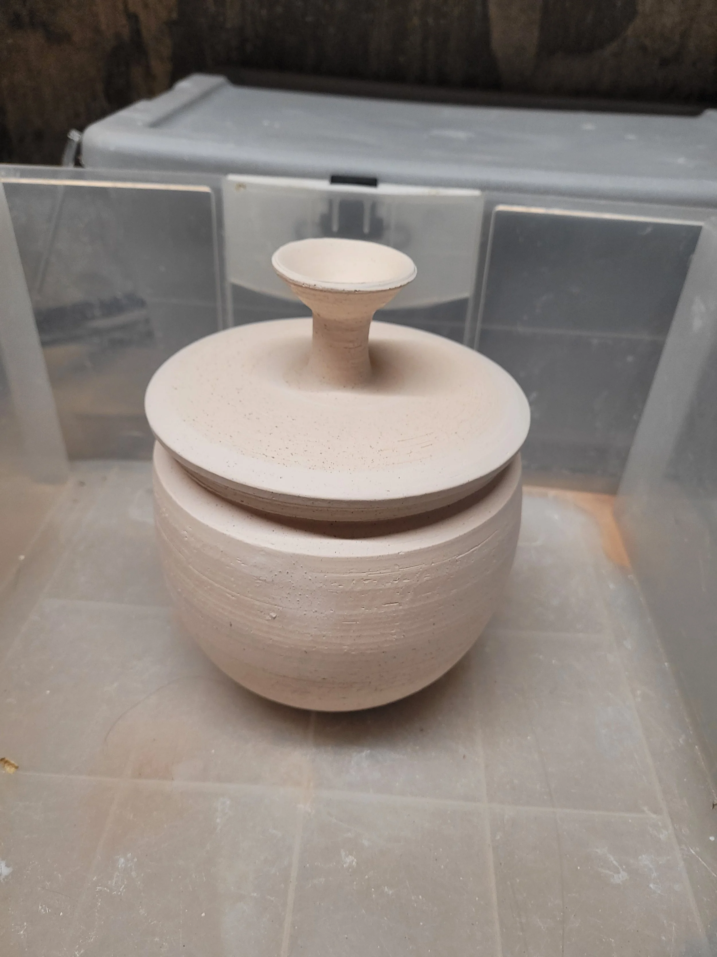 A ceramic vase with a round base and a small, flared opening on top. It is placed on a flat surface inside a display case, with blurred background and gray tiled floor.