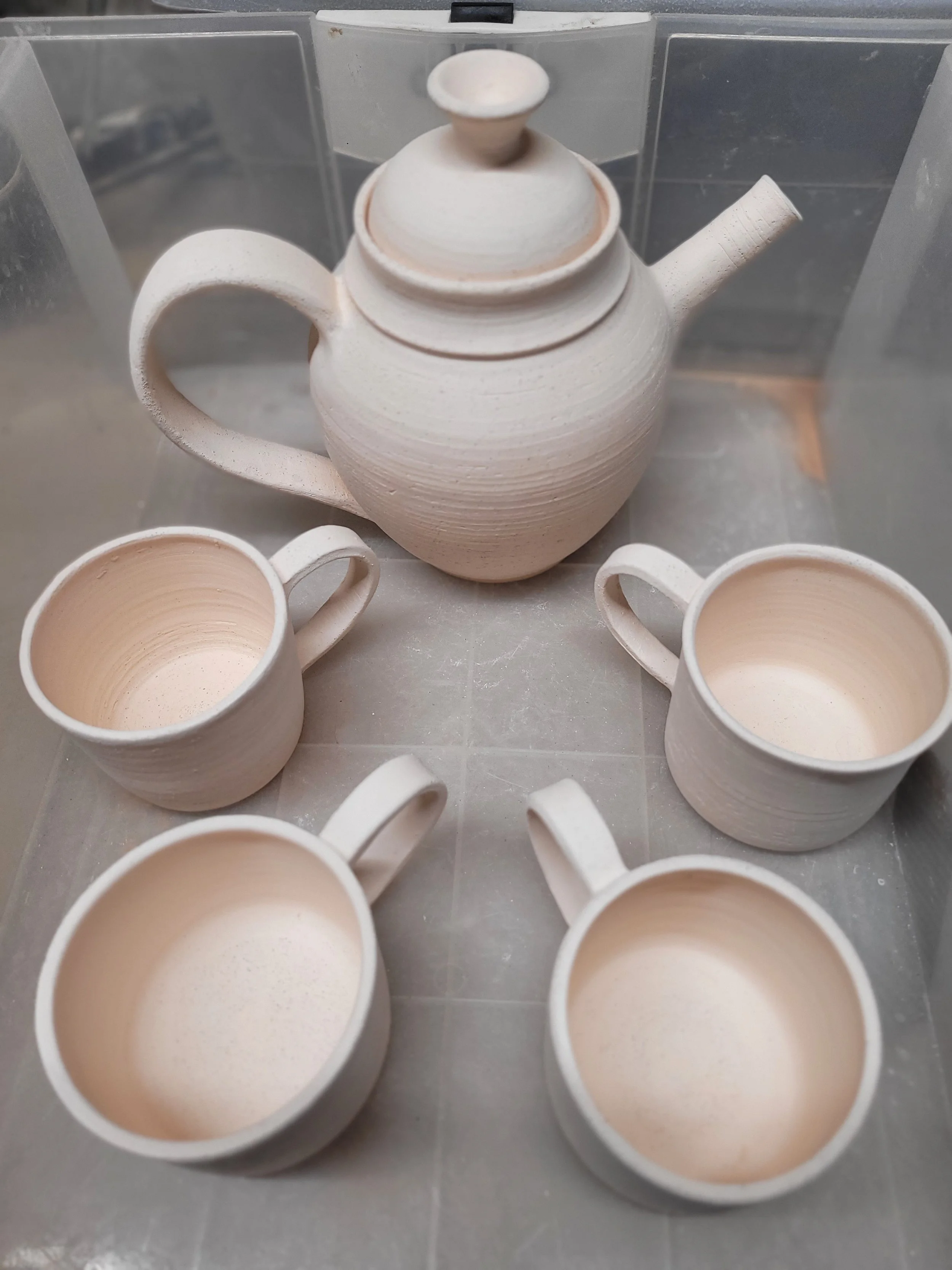 A white ceramic teapot with a round handle and a short, slightly curved spout, positioned among several white ceramic teacups with rounded handles, all inside a transparent plastic storage container.