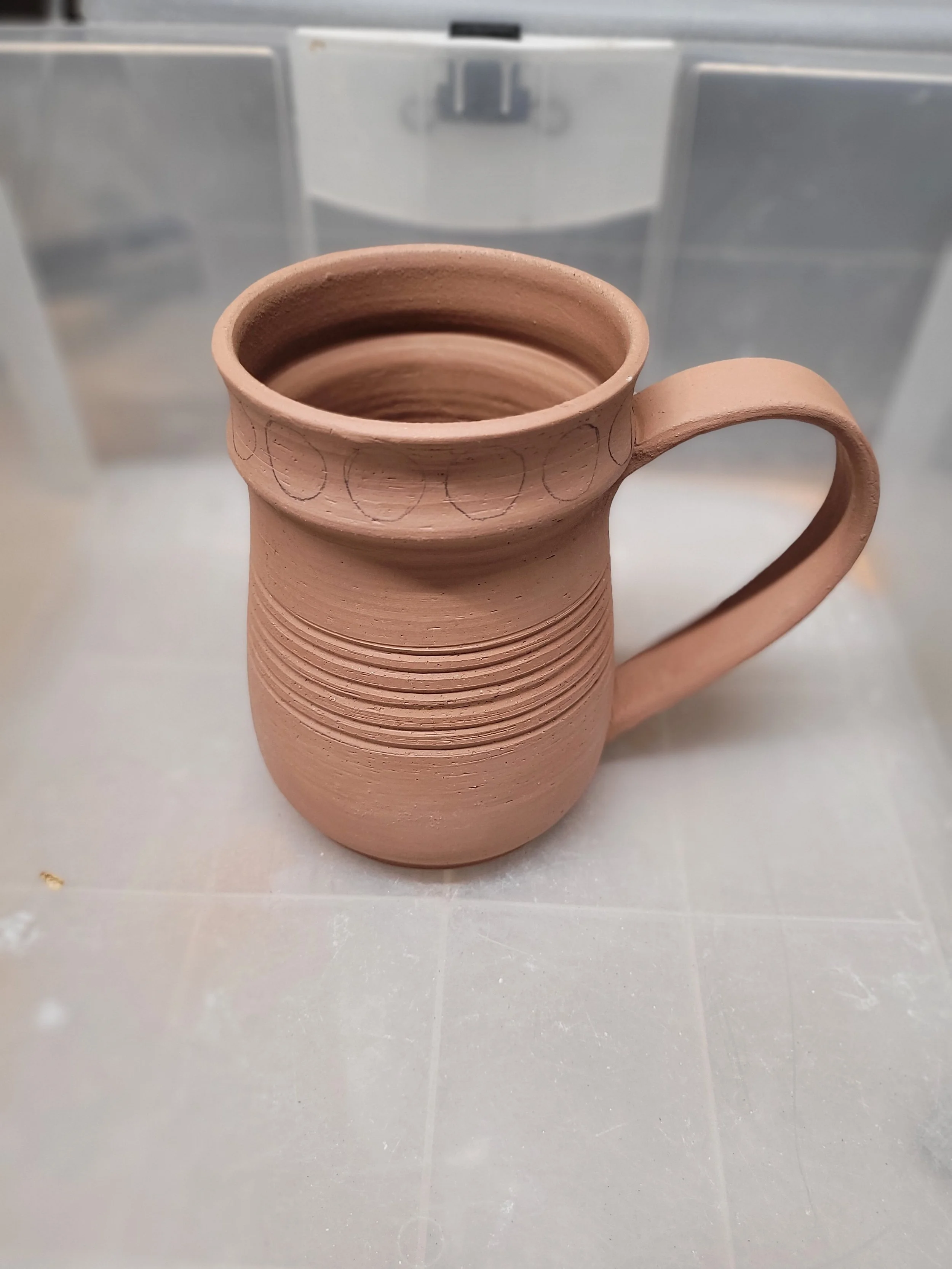 Unfinished clay mug with a handle and circular patterns near the top, placed on a gray surface.