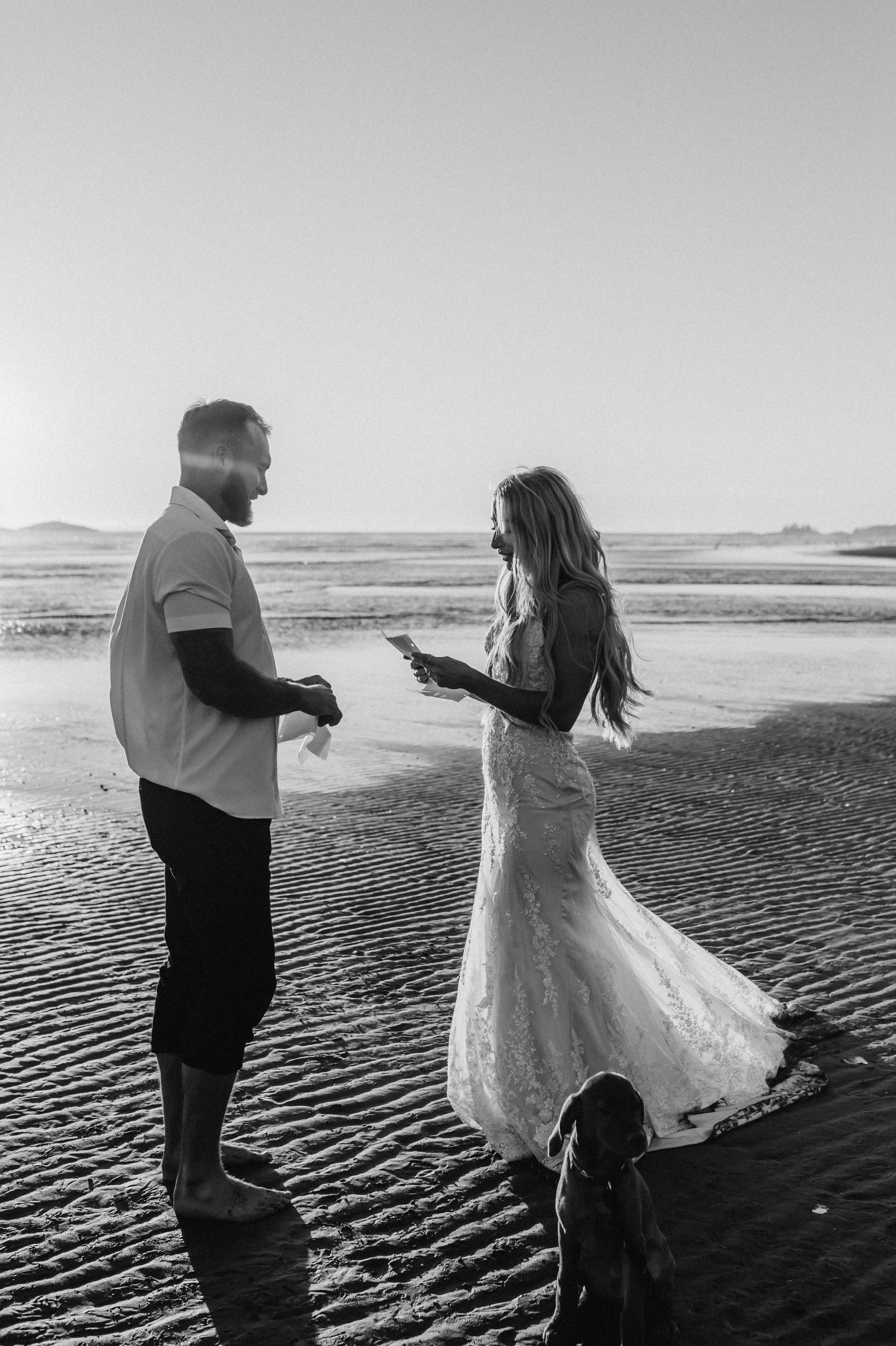 A black and white photo of a man and woman on the beach. They are looking at each other, and a dog is sitting on the sand nearby. The background shows a vast, flat beach with water and distant land. It's their wedding day at Long Beach in Tofino B.C.