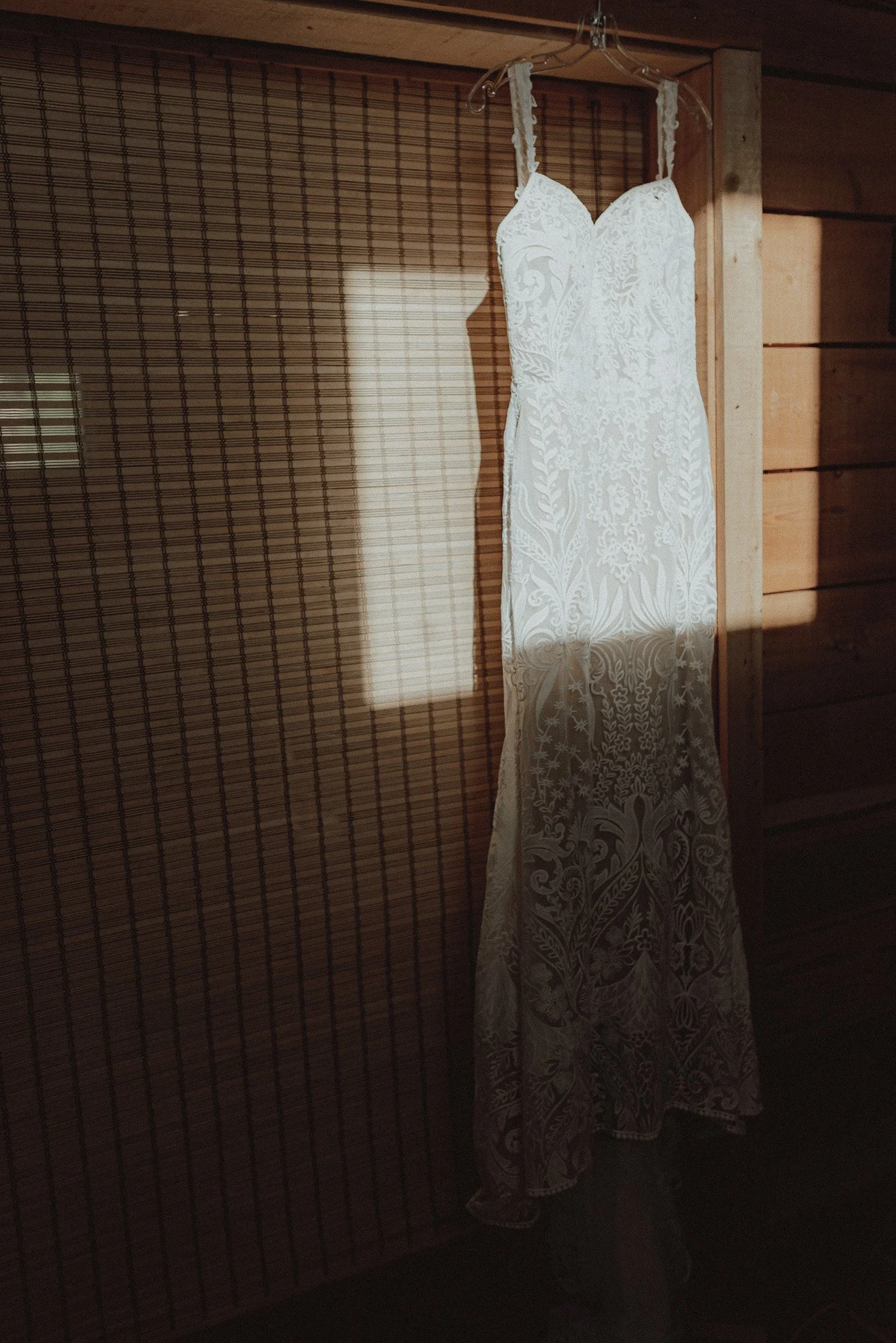 White wedding dress hanging on a hanger against a wooden wall with sunlight creating a shadow.