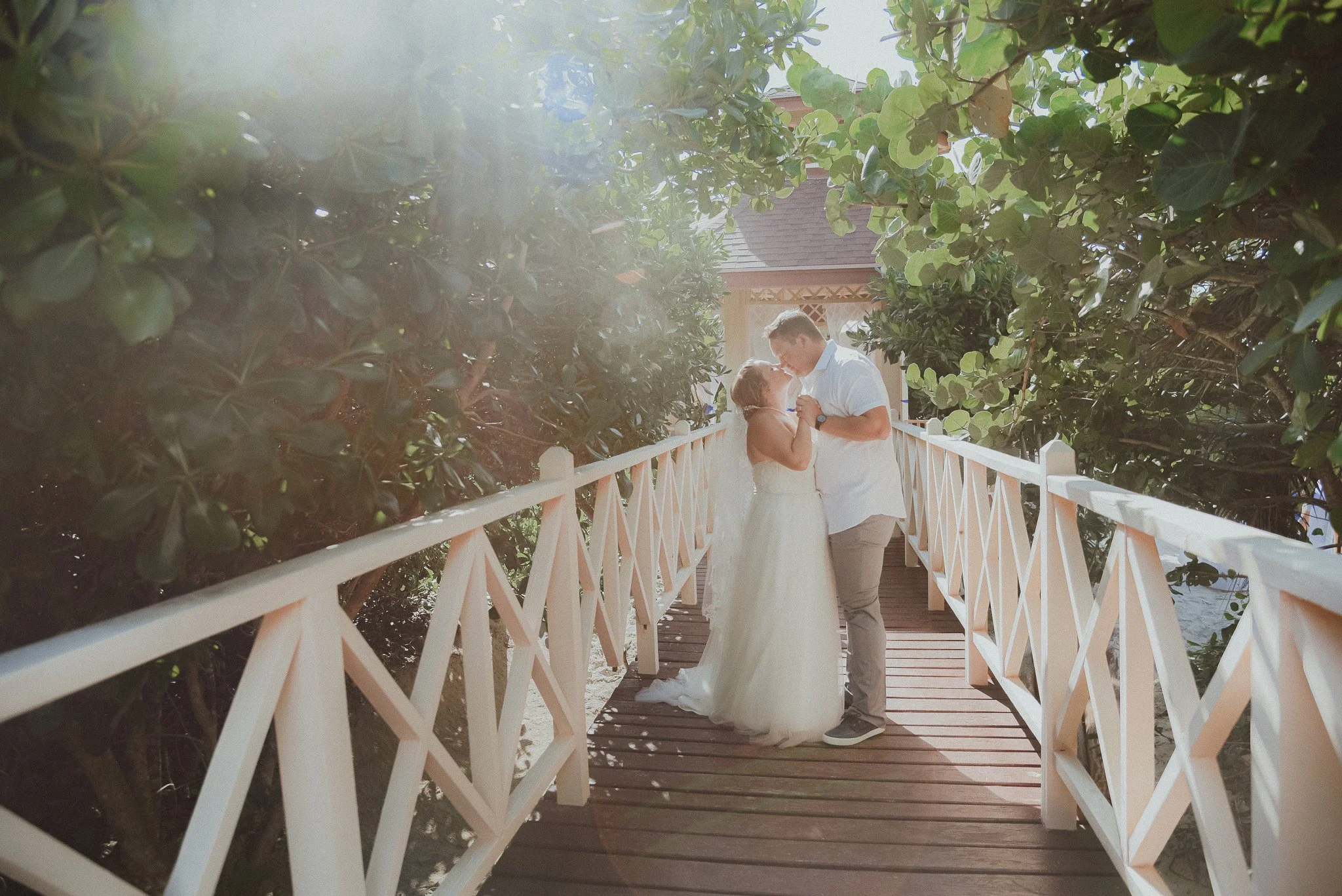 A couple dressed in wedding attire standing close on a small wooden bridge, holding hands, surrounded by lush green foliage with sunlight filtering through. Veradero Cuba