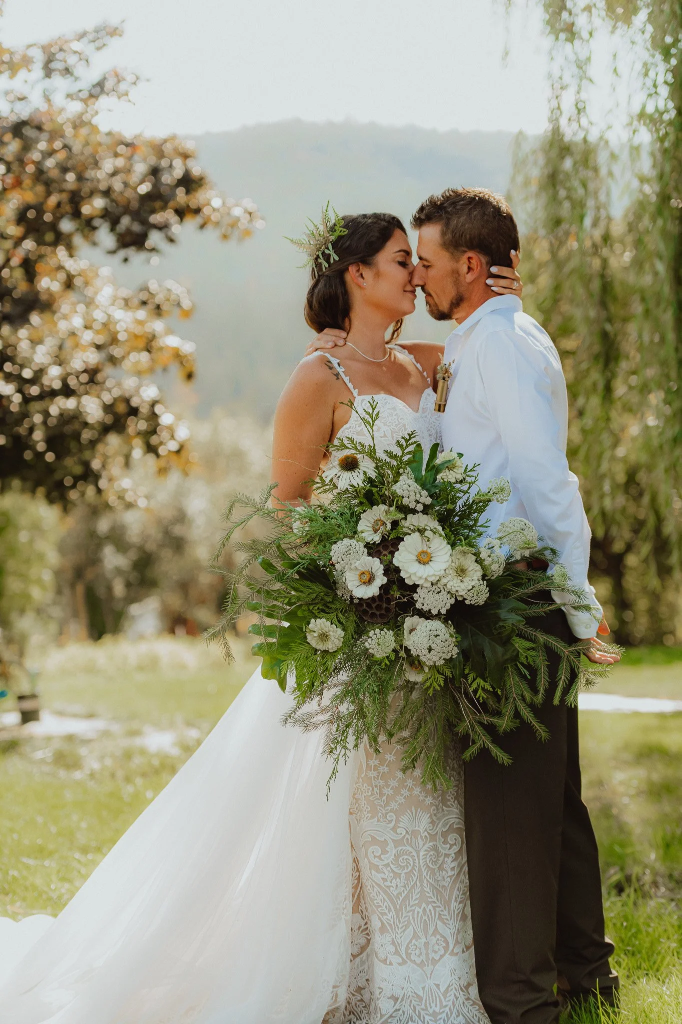 A bride and groom sharing a close moment outdoors, the bride holding a large bouquet of white flowers and greenery, with trees and mountains in the background. Vernon B.C.