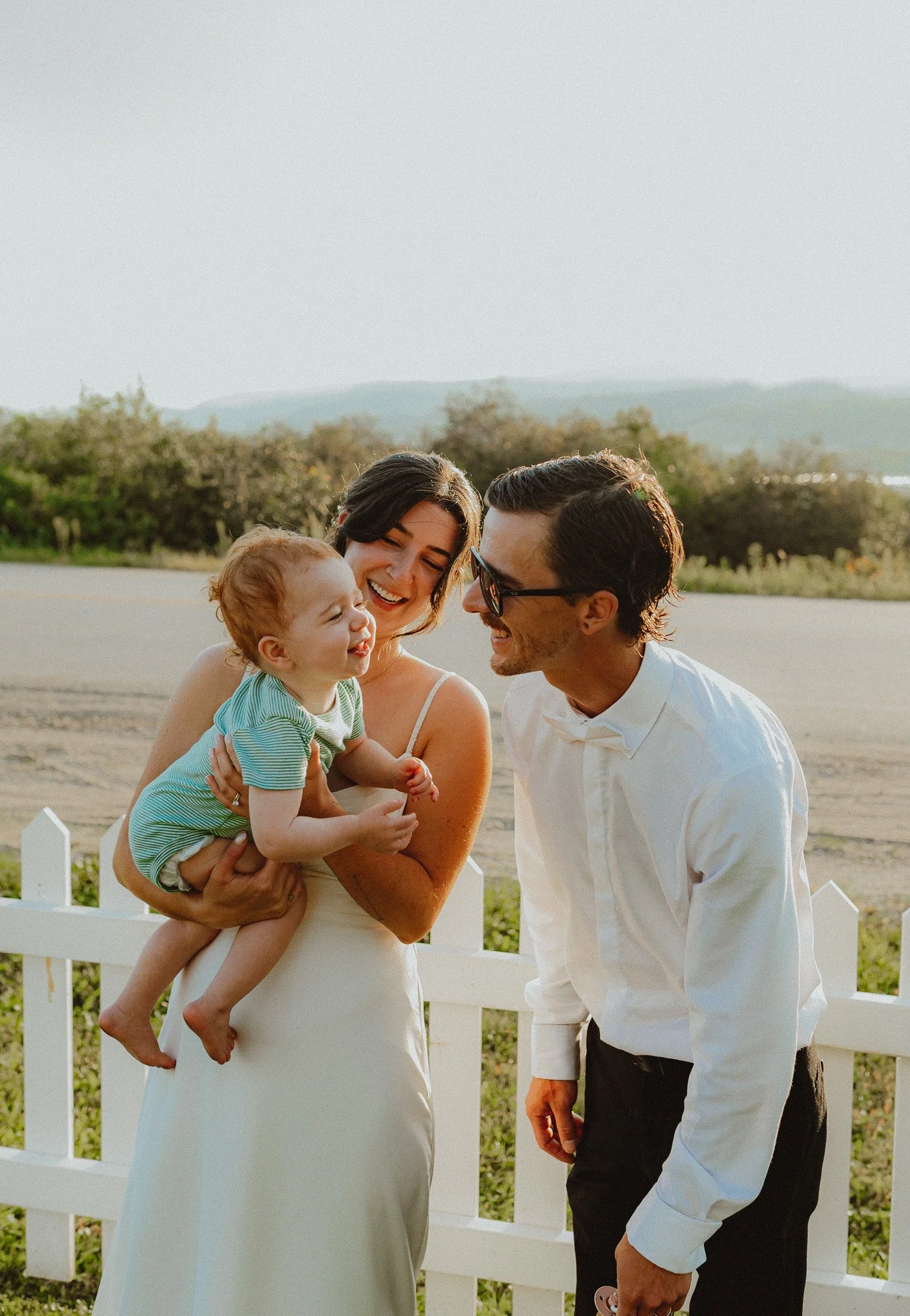 A smiling woman holding a young child in her arms with a man leaning in, all outdoors near a white wooden fence, during golden hour with a landscape of trees and hills in the background.