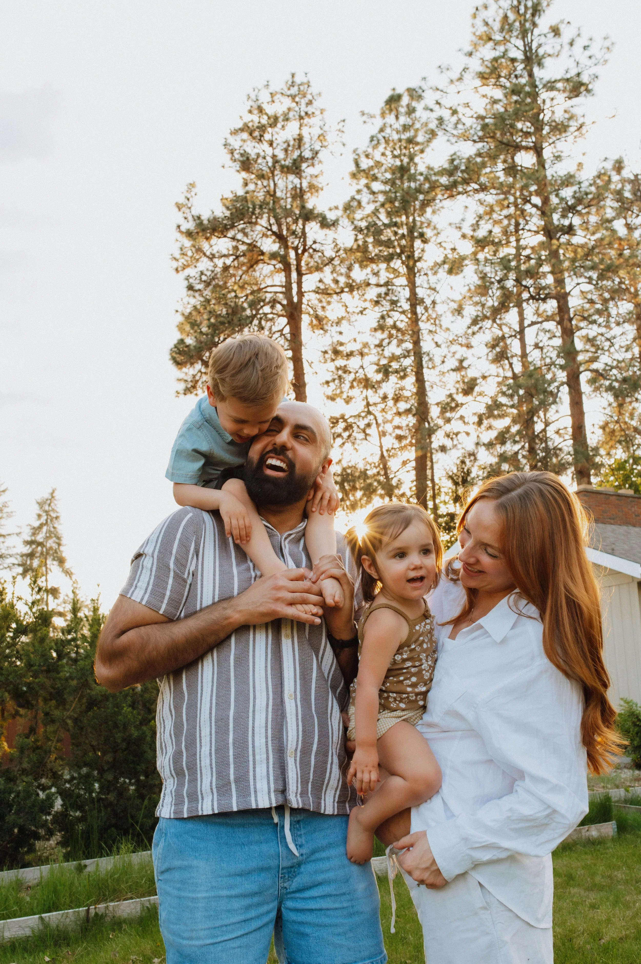 Family of four outdoors during sunset, with tall trees in the background. The man with a beard is holding a young boy on his shoulders, while a woman with long red hair holds a young girl, all smiling and enjoying the moment.