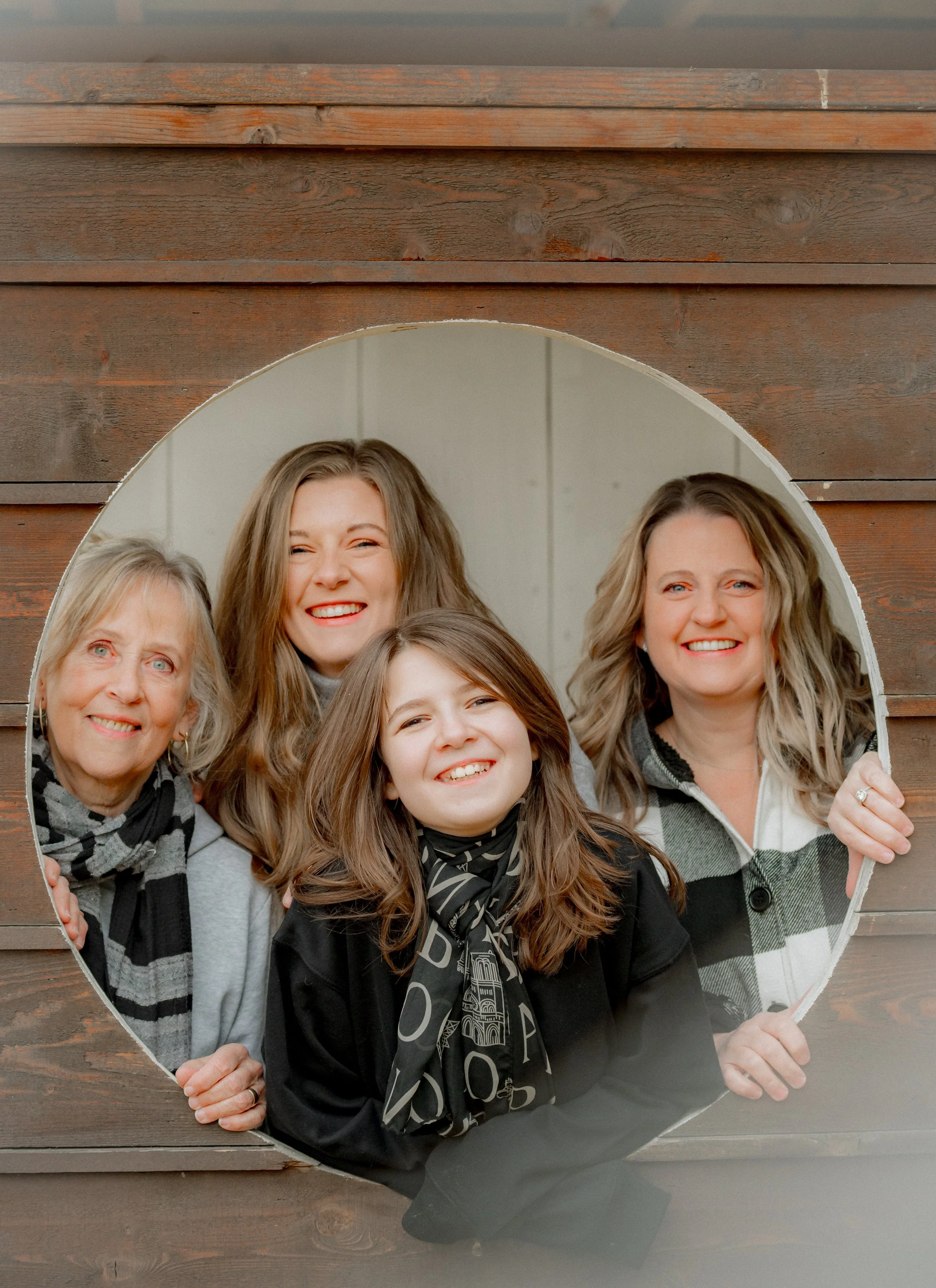 Four women and one girl smiling through a circular cutout in a wooden wall.