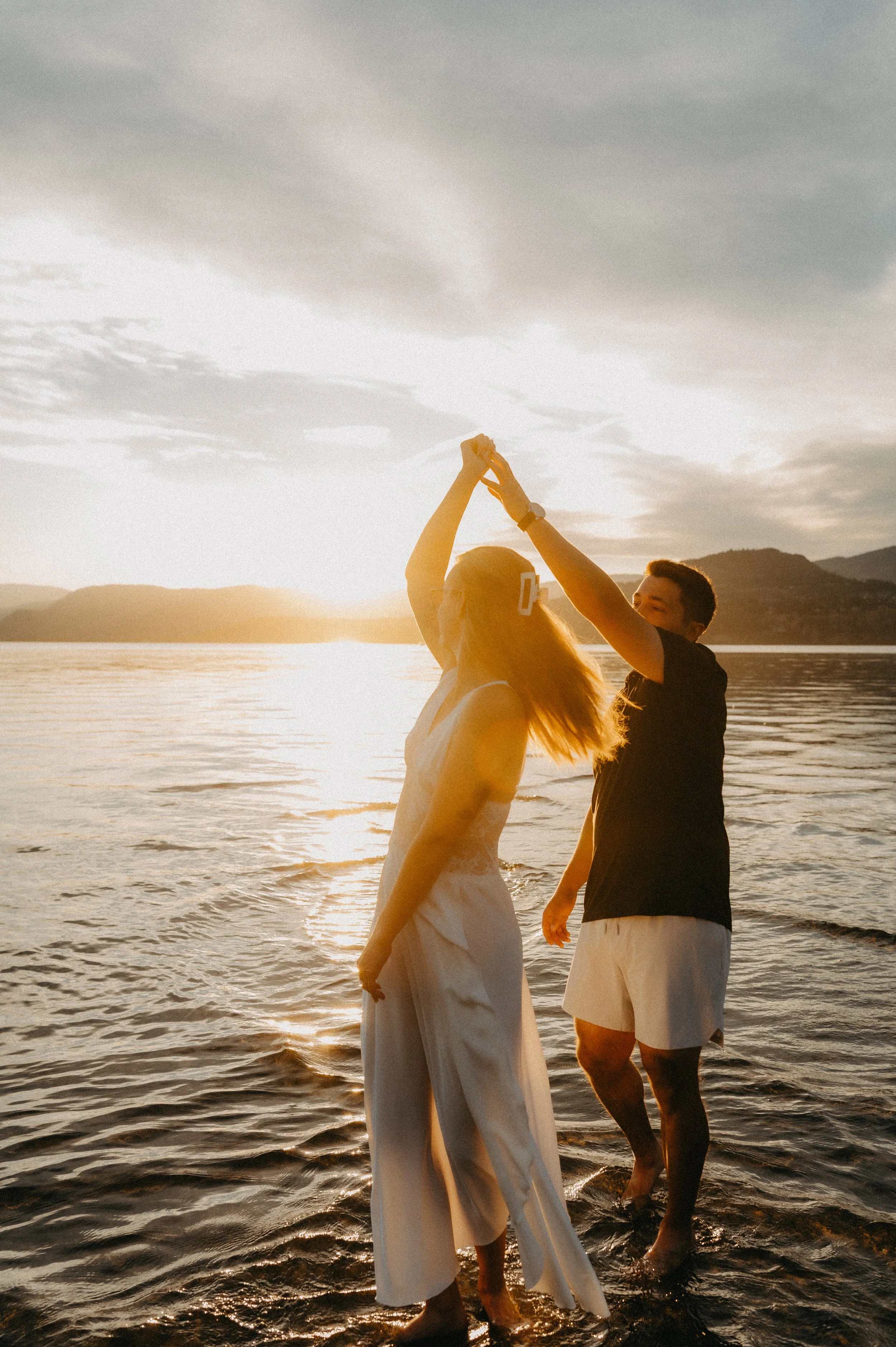 A couple dancing in the water during sunset, holding hands above their heads, with mountains in the background. Okanagan Lake, Kelowna B.C.