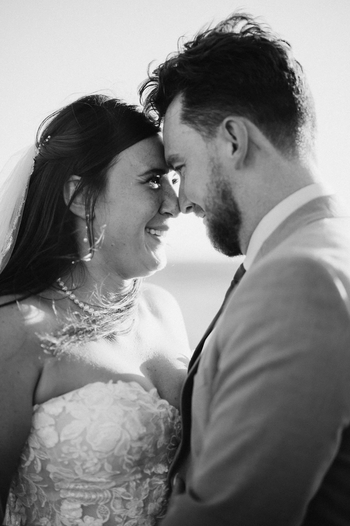 A black and white photo of a bride and groom with foreheads touching, smiling at each other during their wedding.