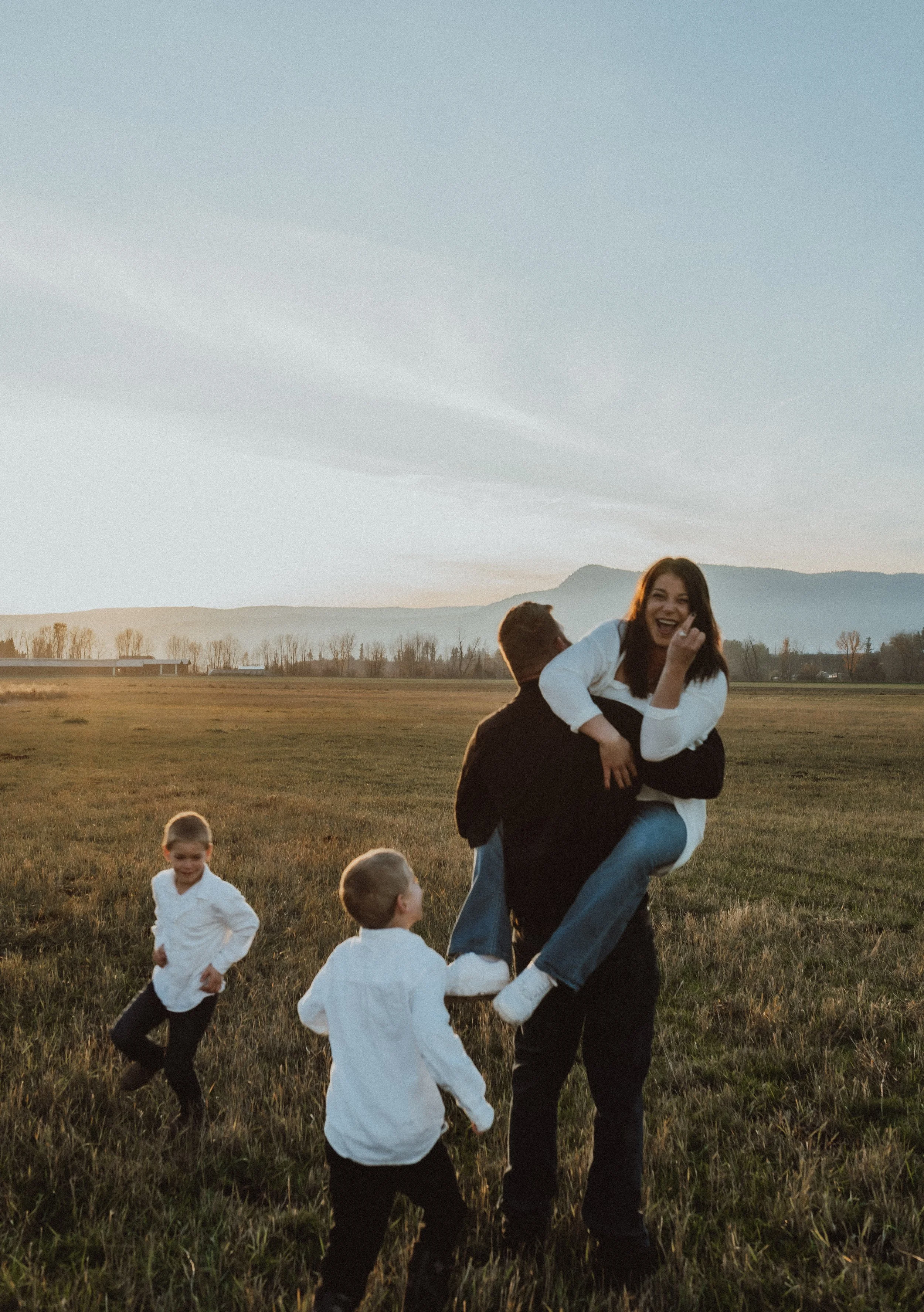 Family of five enjoying outdoors during sunset, with a woman being lifted by a man, all smiling, children playing nearby in a grassy field with mountains in the background.