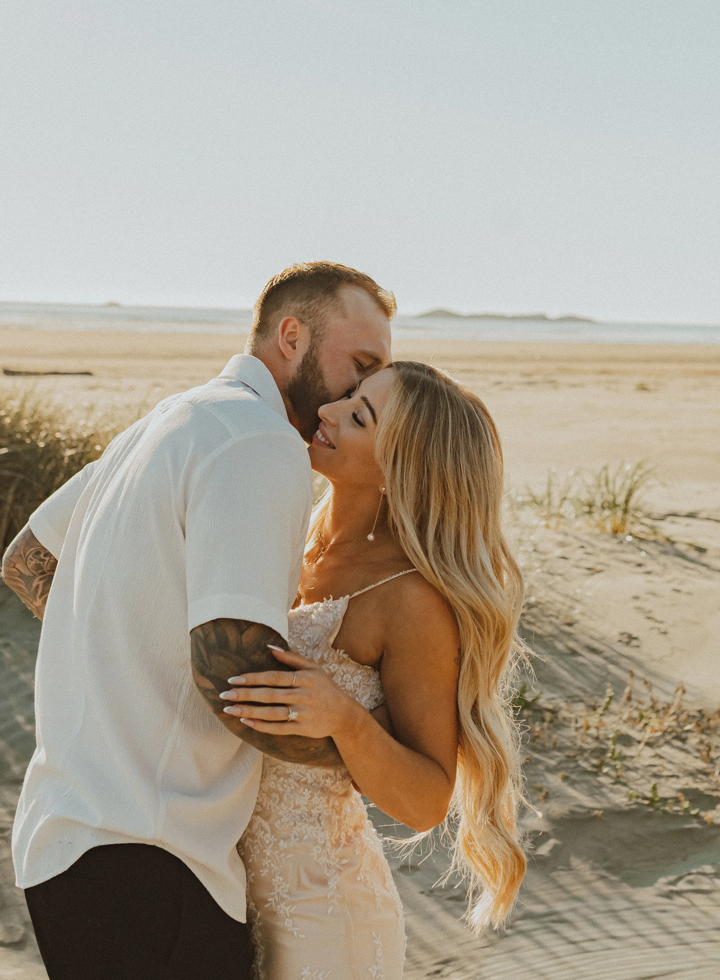 A couple kissing on a beach at sunset, dressed in wedding attire with the man in a white shirt and the woman in a lace wedding dress.