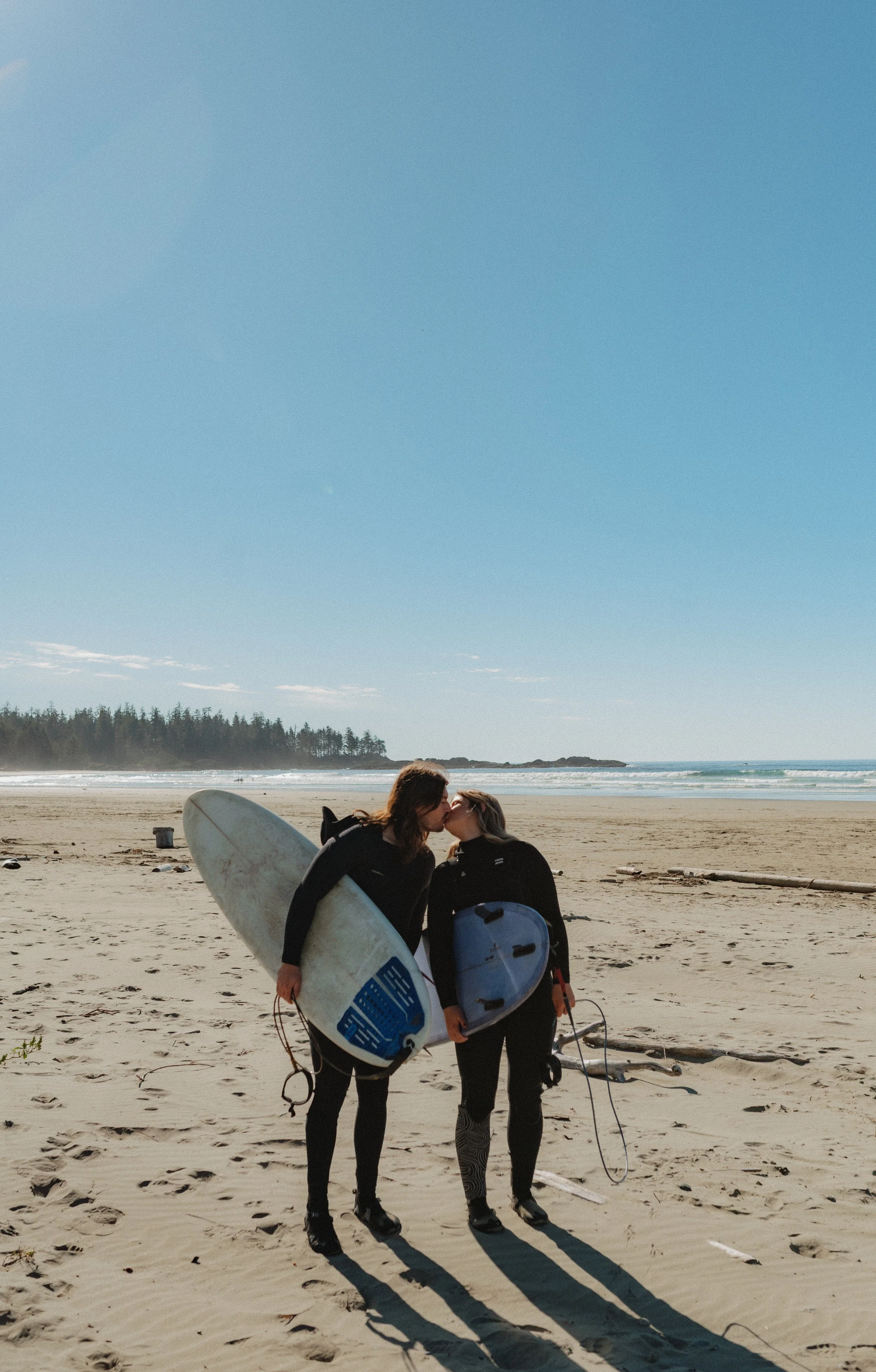 Two women in wetsuits share a kiss on a sandy beach, holding surfboards. The ocean and a tree-lined coastline are visible in the background.