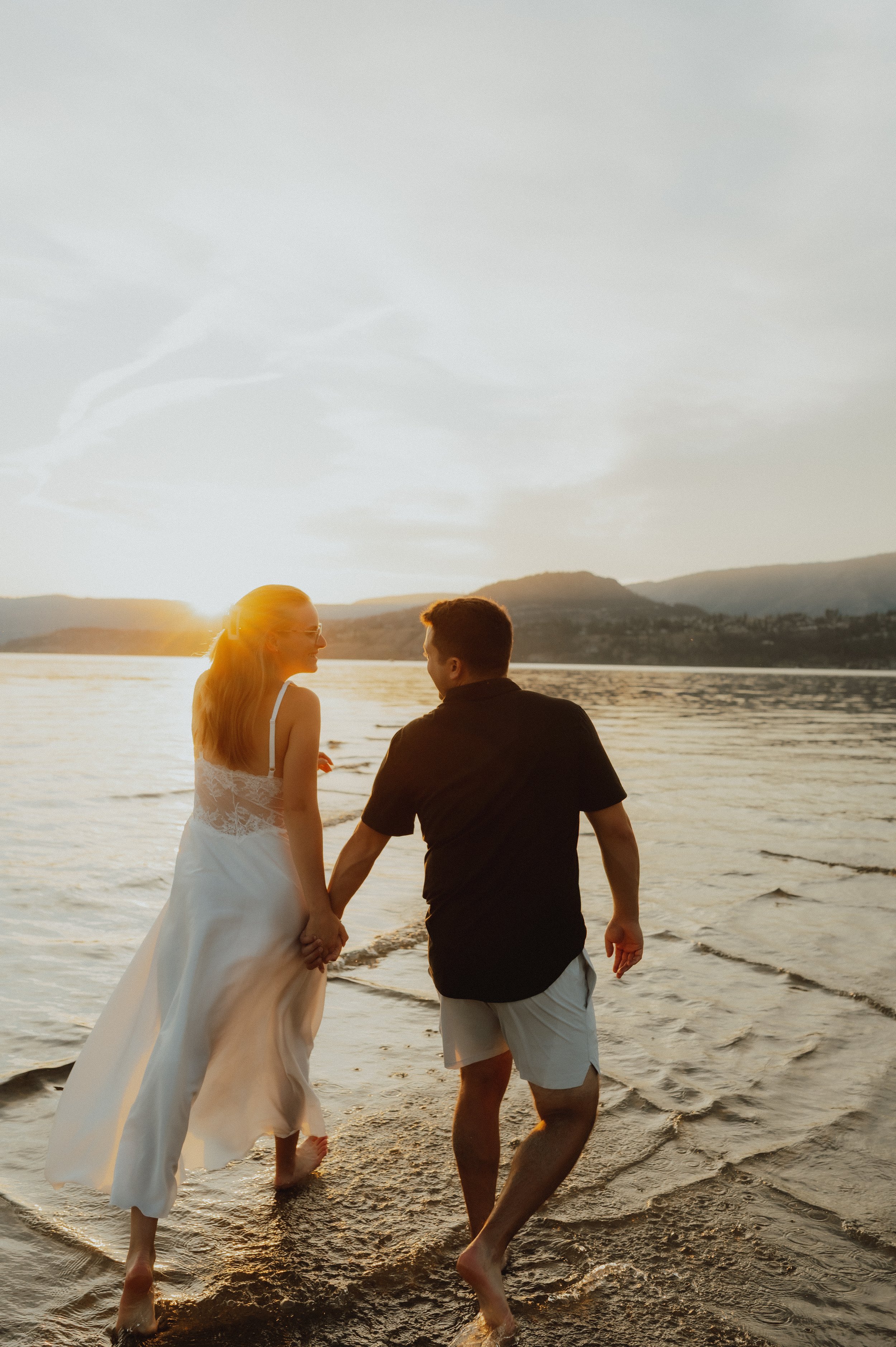 A couple holding hands while walking along the shoreline at sunset, with the sun setting behind distant mountains.