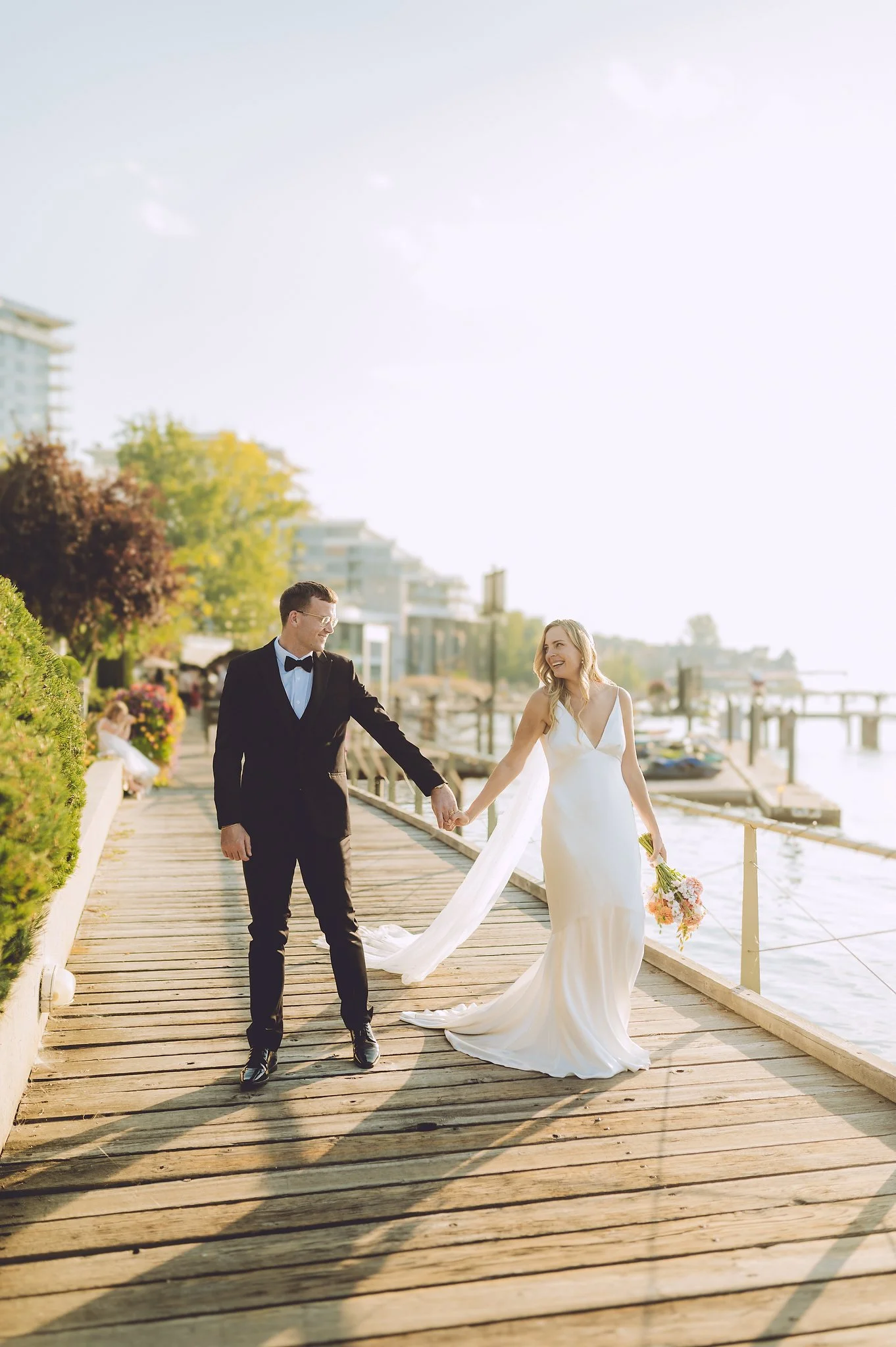 A bride in a white wedding dress and a groom in a black tuxedo walking hand-in-hand on a wooden pier by the water, holding a bouquet of flowers, with buildings and trees in the background.  Manteo Resort, Kelowna B.C.