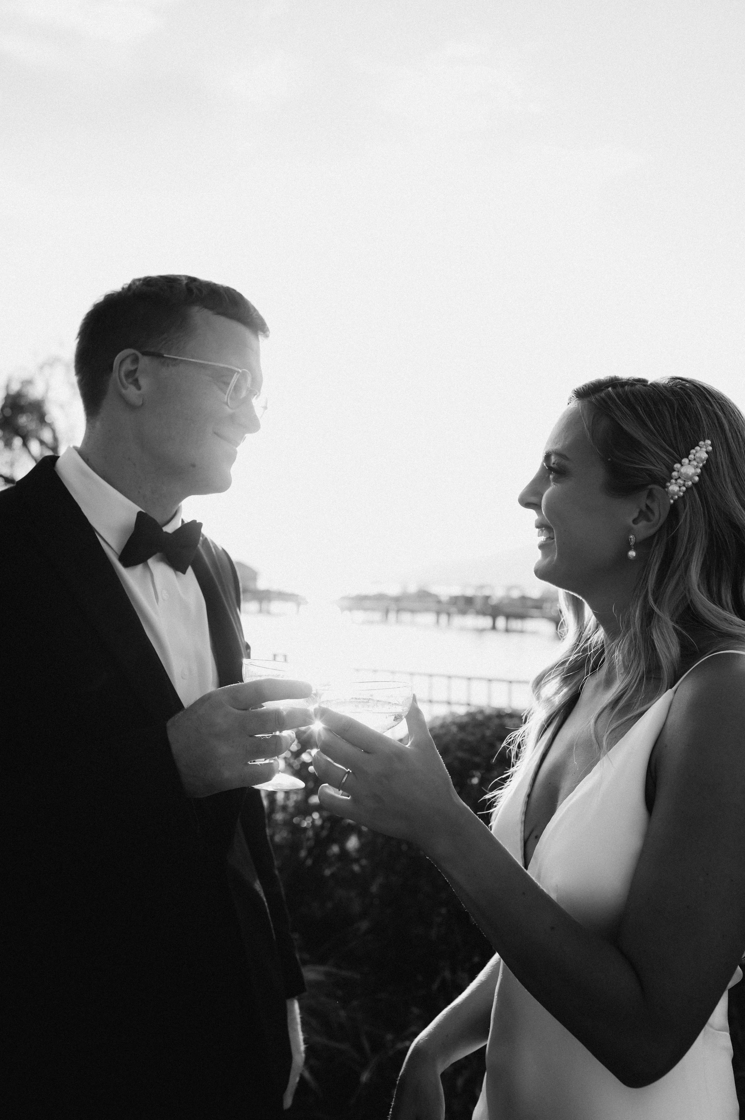 A black and white photo of a wedding couple outdoors, smiling and holding glasses in a toast with a body of water and pier in the background.  Manteo Resort, Kelowna B.C.