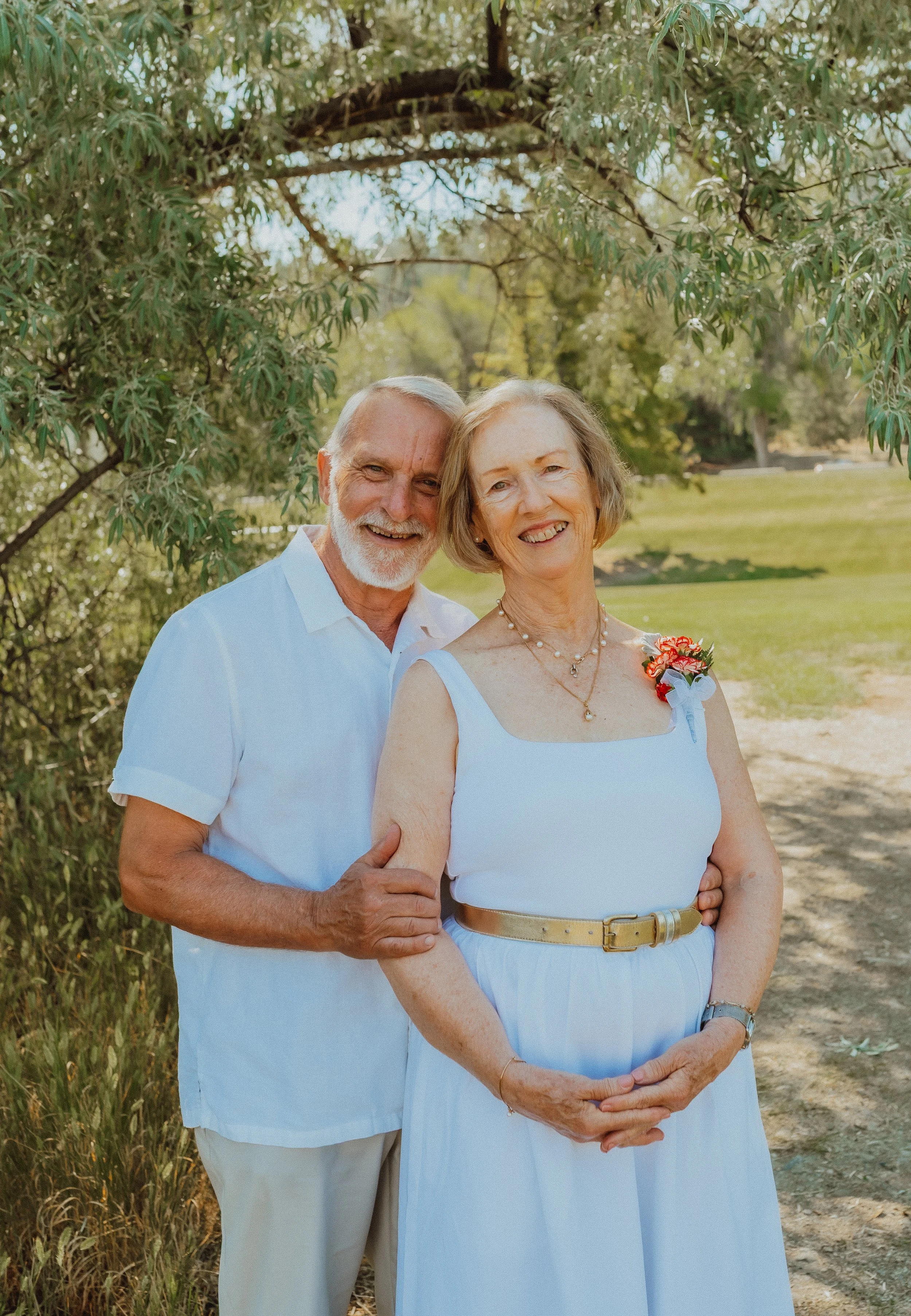 An elderly couple standing outdoors under trees, smiling. The woman is wearing a white sleeveless dress with a beige belt and a floral boutonniere, while the man is in a white short-sleeved shirt and beige pants.