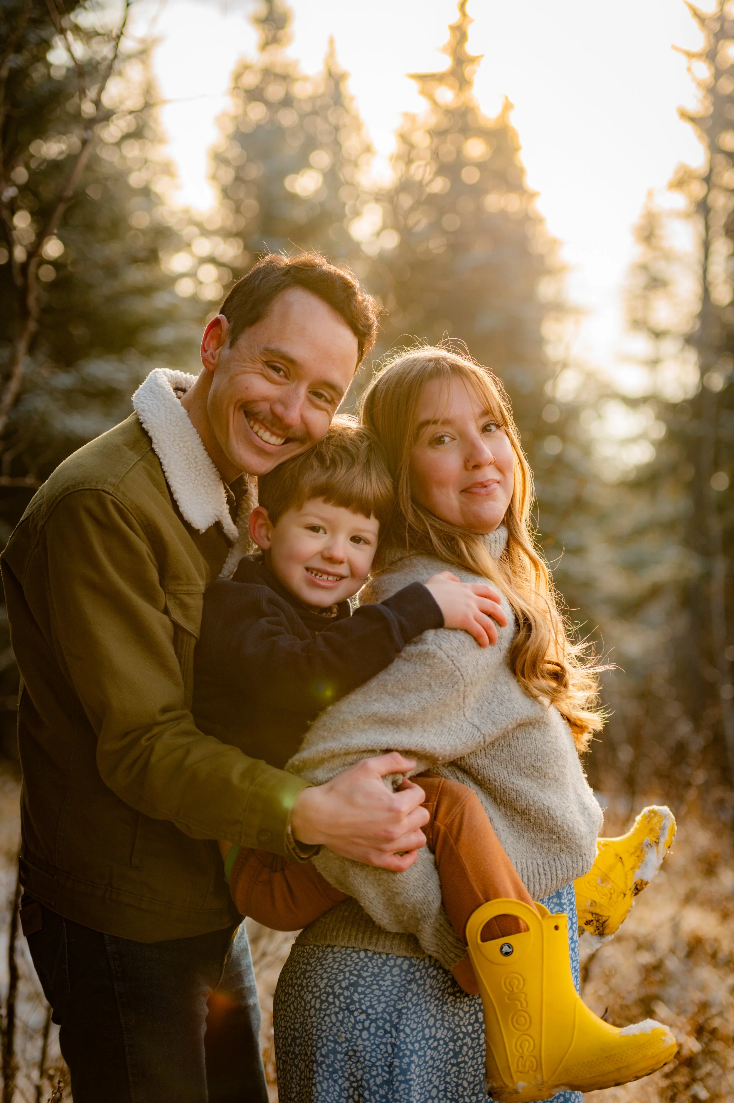 Family of three in the woods enjoying sunset, the man has brown hair and is wearing a brown jacket with a shearling collar, the woman has long red hair and is wearing a gray sweater, the child has brown hair and is wearing a black jacket and yellow r