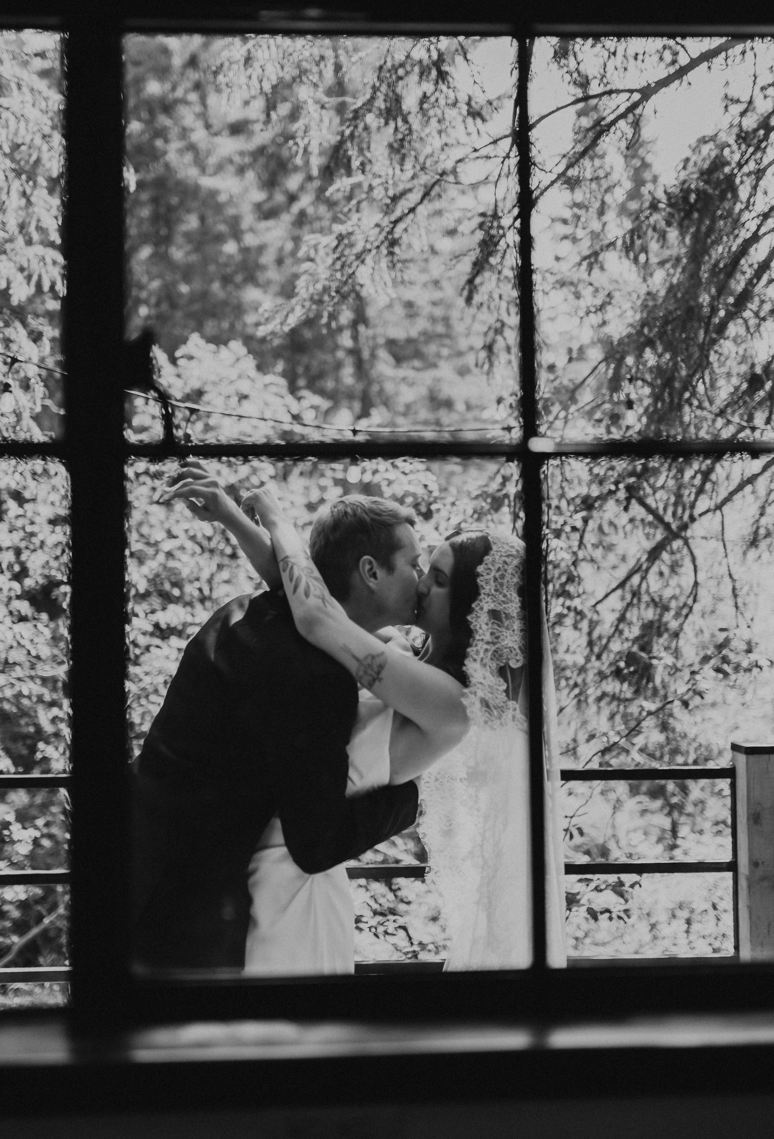 A black and white photo of a couple kissing outside, viewed through a window with a grid of panes, with trees in the background. The Valley Wedding Venue, Westerose Alberta