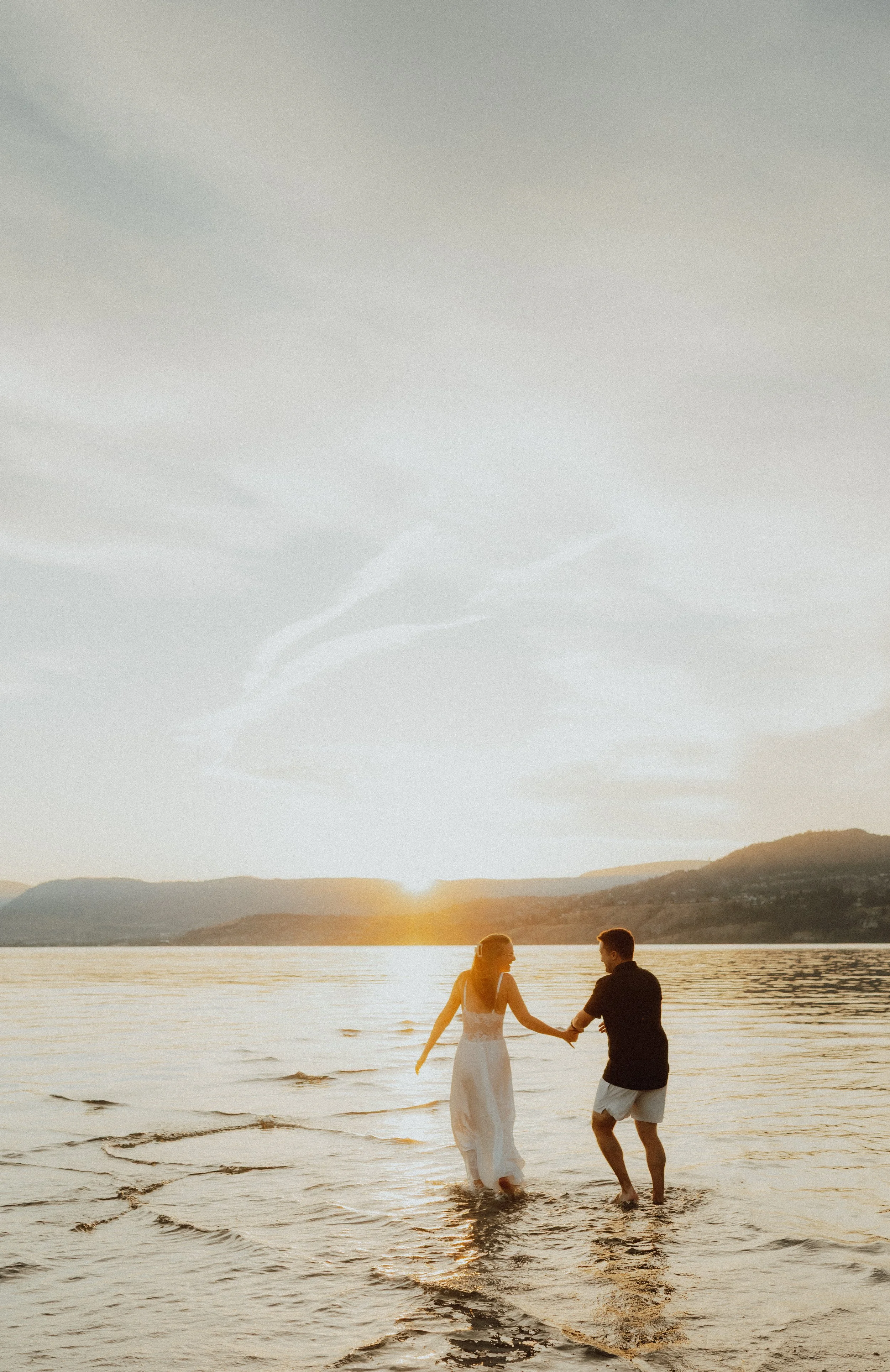 A couple holding hands and walking in the water at sunset, with mountains in the background.