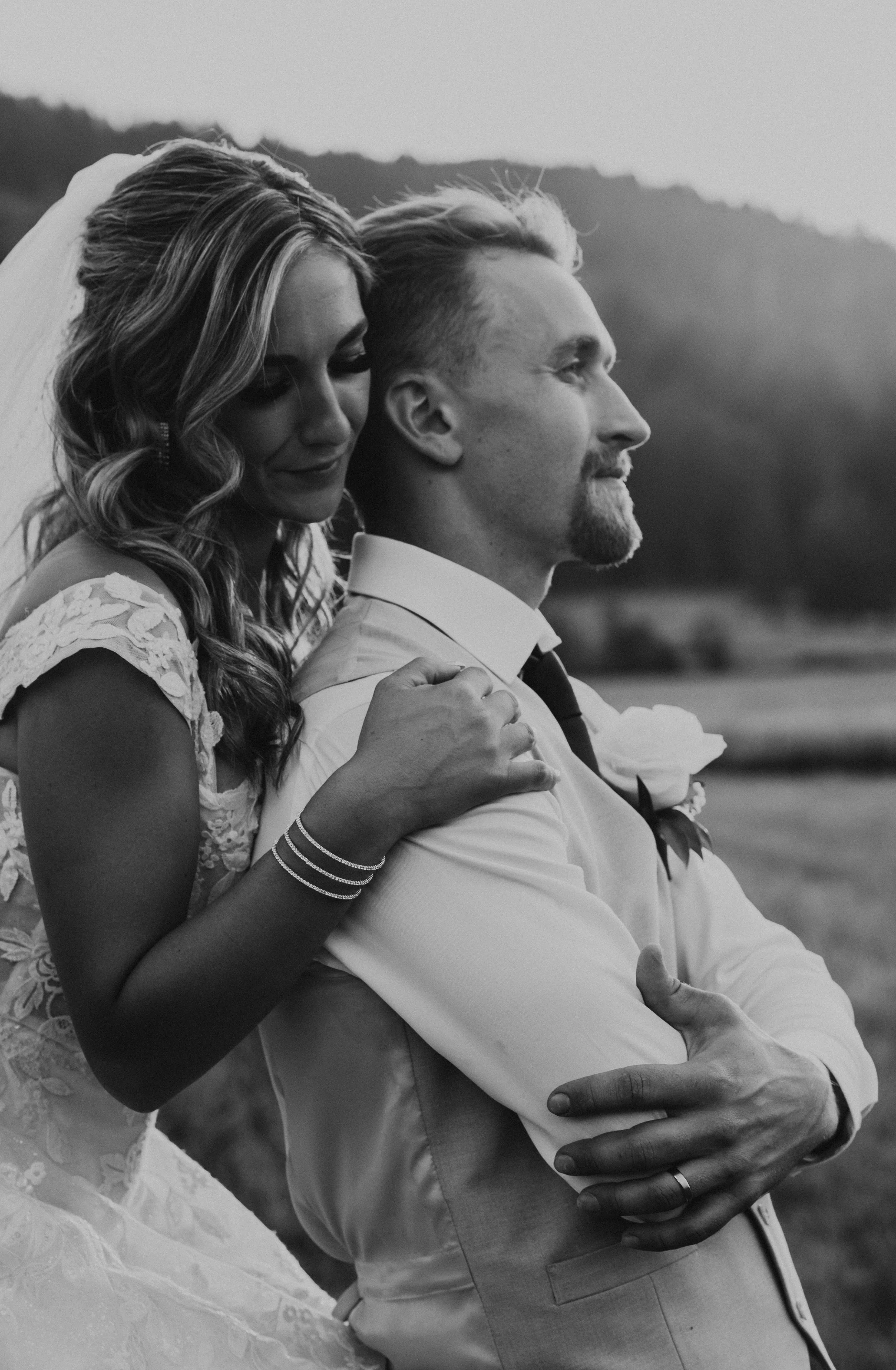 A black and white photo of a bride and groom outdoors, with the bride hugging the groom from behind, both showing happy expressions.