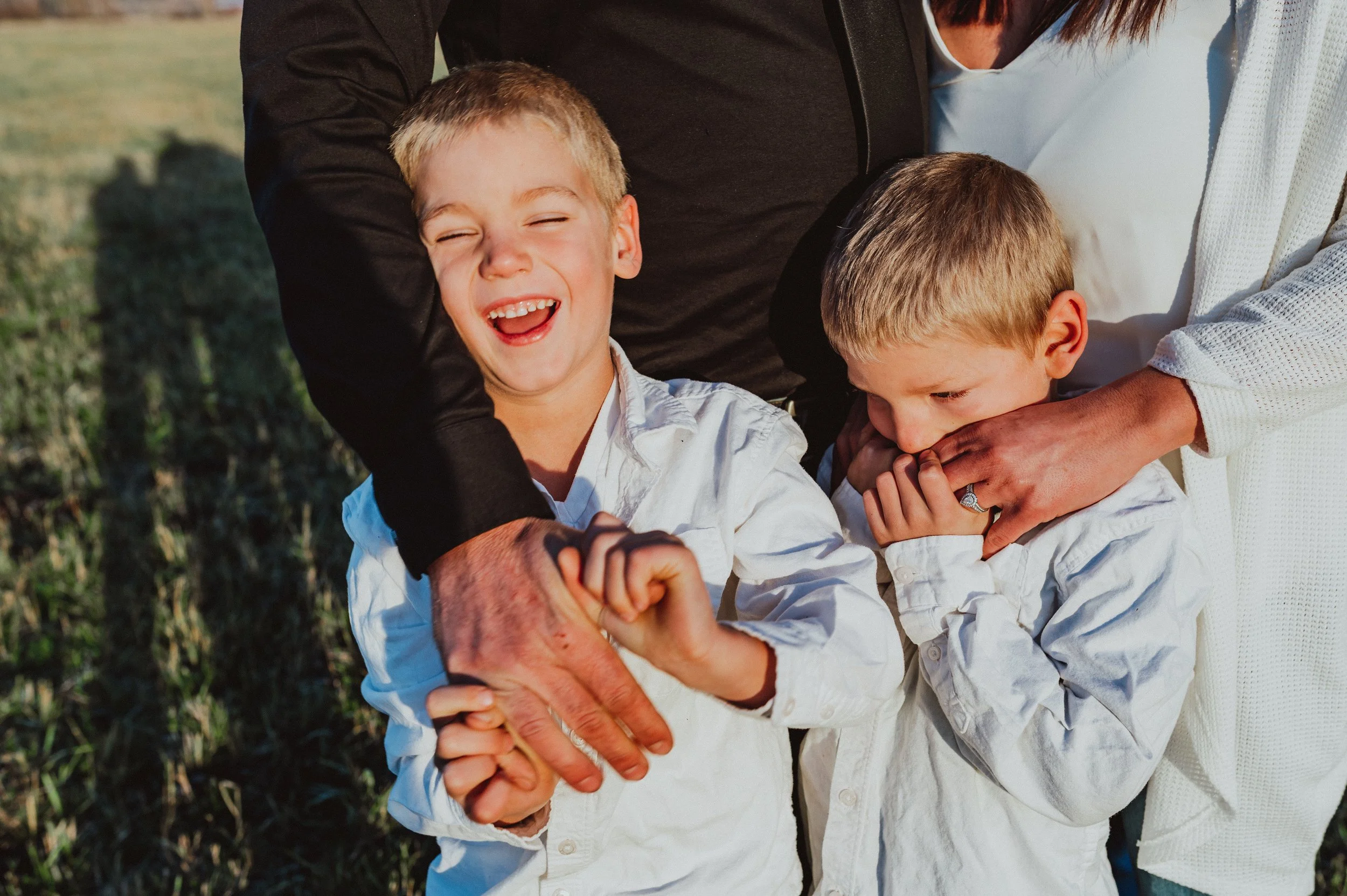A joyful family moment outdoors with two young boys and their parents. The older boy is smiling and laughing while holding hands with an adult. The younger boy appears shy or coy, being gently held by an adult. All family members are wearing white shirts, with the background showing a grassy field.