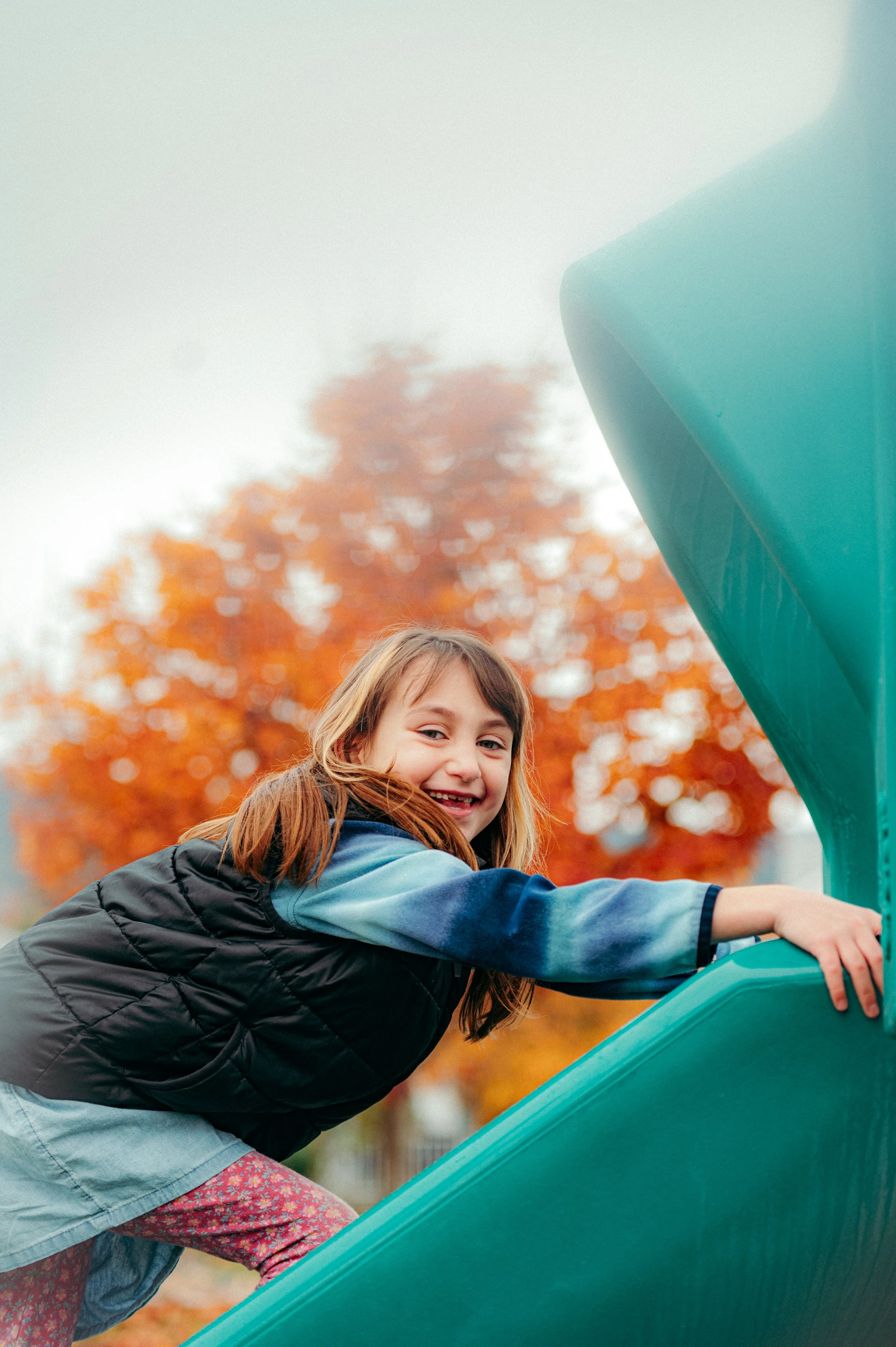 A young girl with light brown hair smiling and holding onto a green slide at a playground during autumn, with orange and red trees in the background.