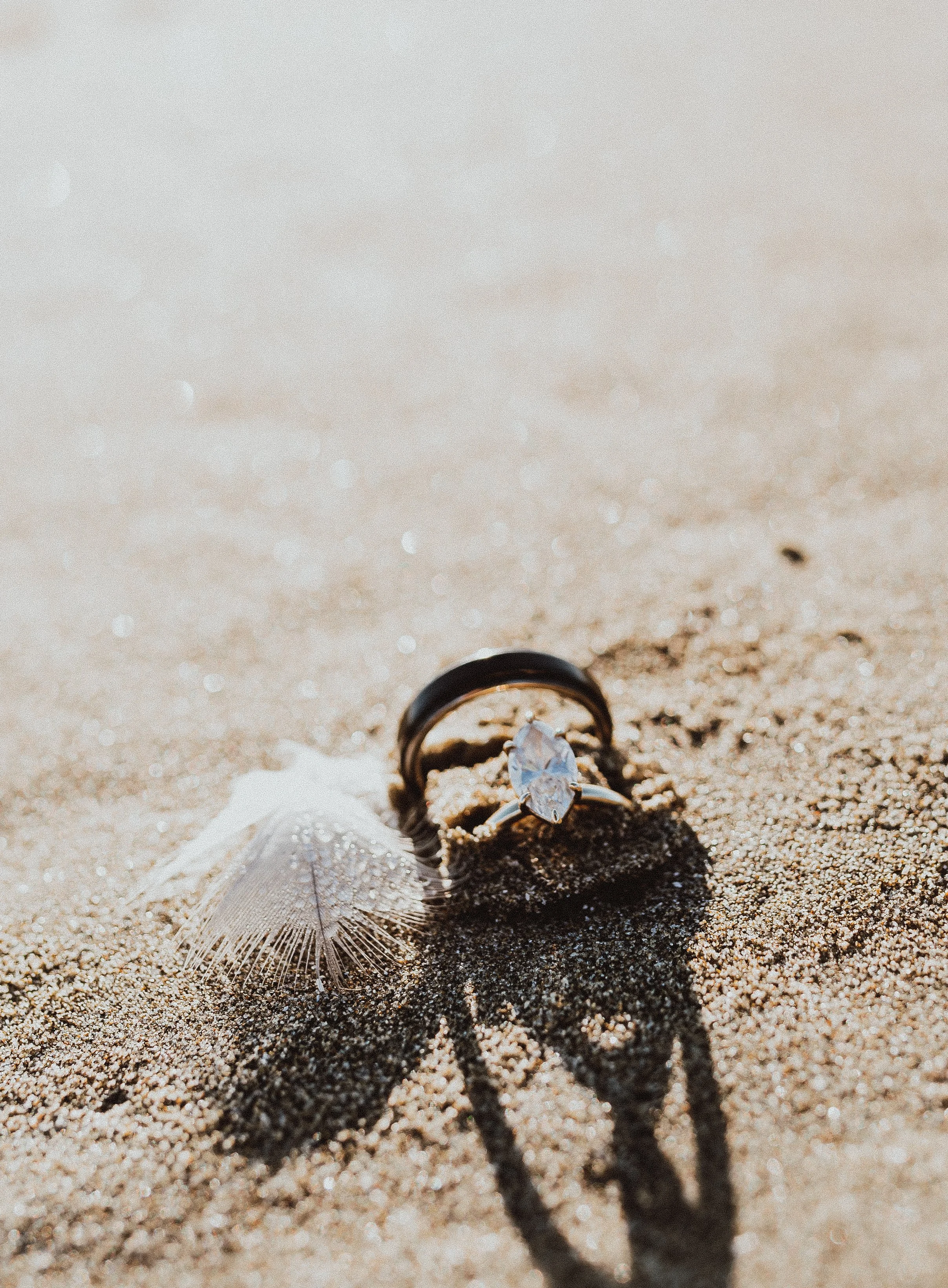 Engagement ring with a large oval diamond, wedding band, shell, and shadow on sandy beach during daytime.