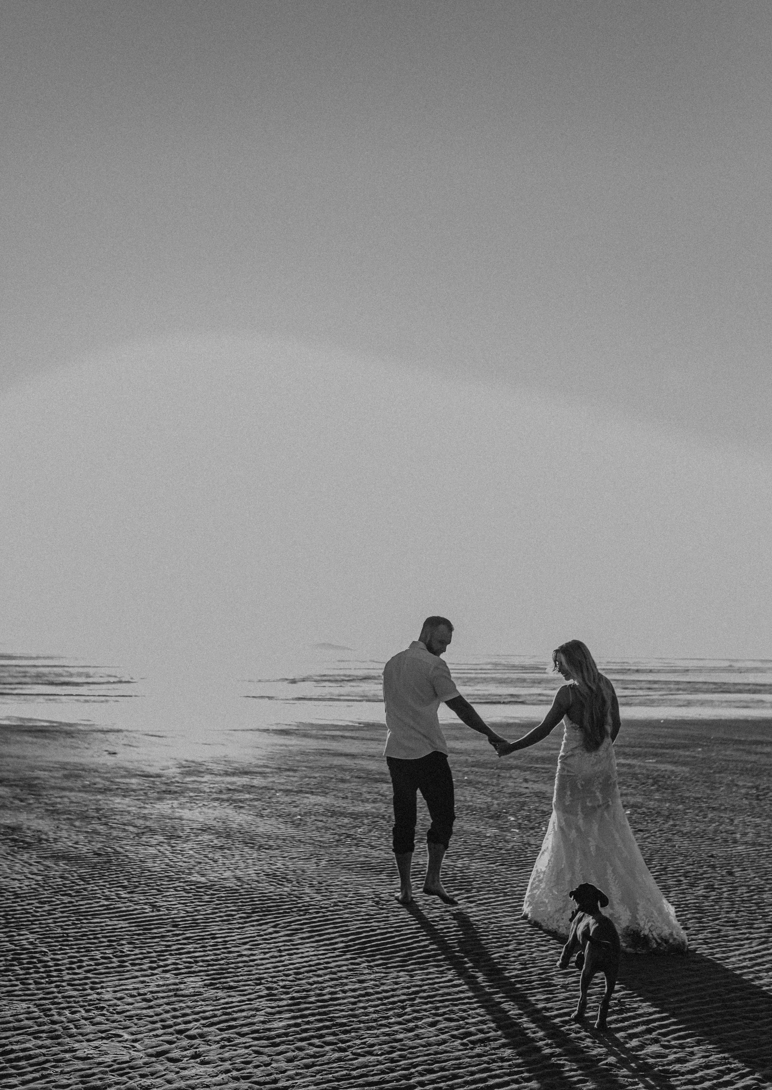 A couple holding hands on a beach at sunset, with a dog nearby.