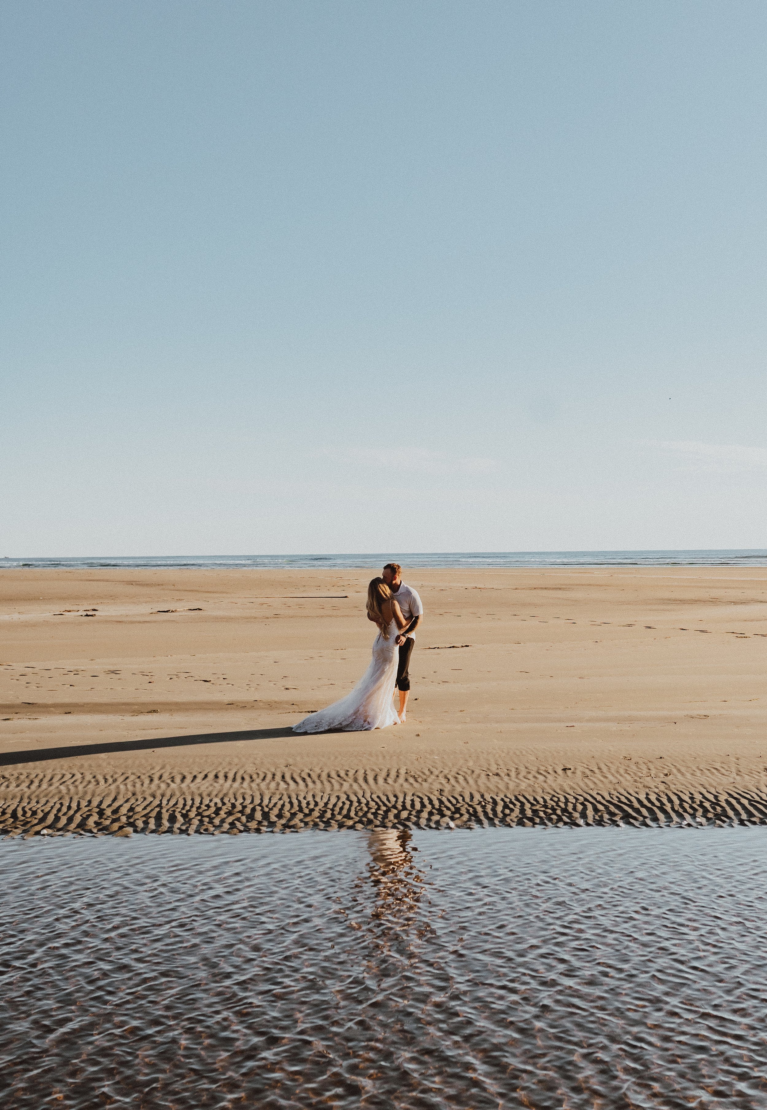 A couple kissing on a sandy beach during sunset, with the ocean in the background and their reflection in shallow water.