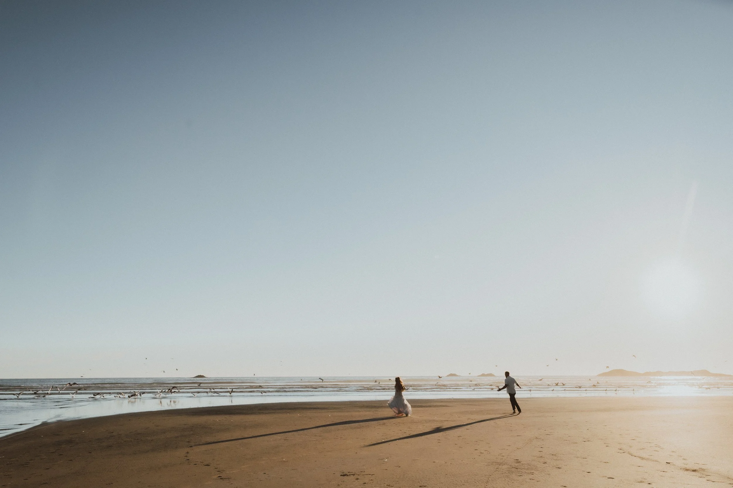 A couple dancing on the beach during sunset, with seagulls flying overhead and distant islands on the horizon.