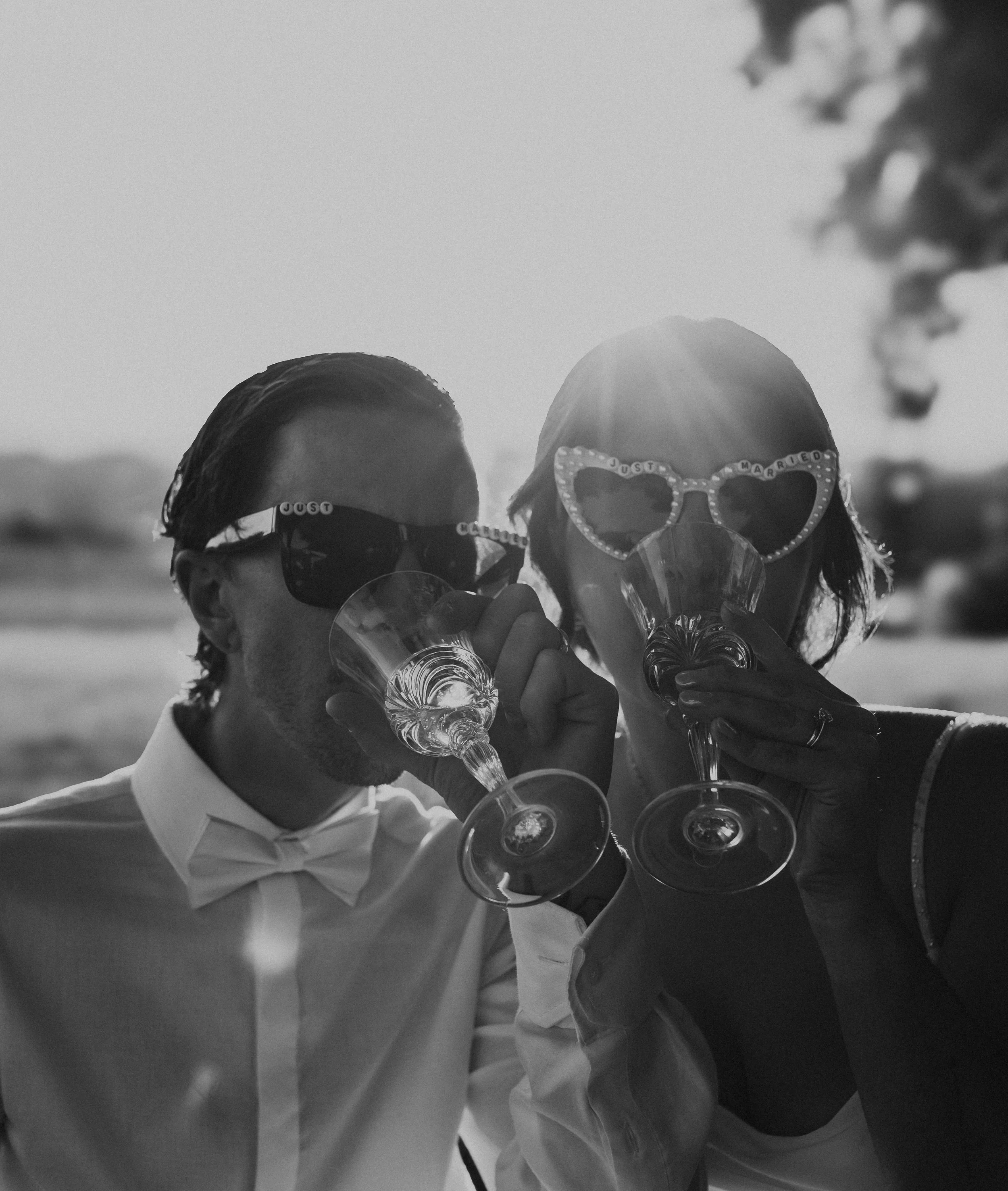 Black and white photo of a couple dressed in wedding attire enjoying drinks outdoors during sunset, both wearing sunglasses and unique heart-shaped glasses, toasting with wine glasses.