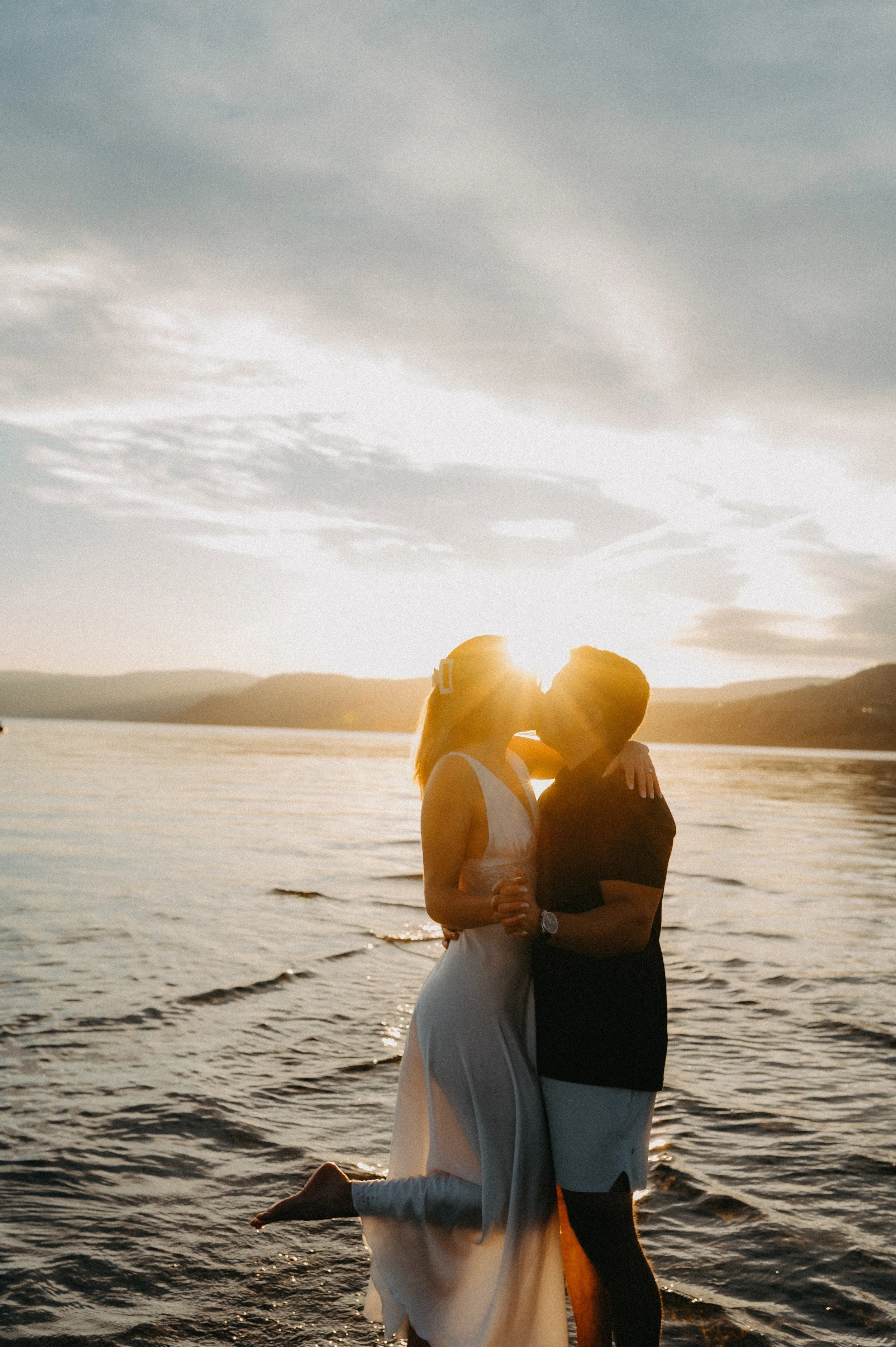 A couple embracing at sunset by the Okanagan Lake, with mountains in the background. Kelowna, B.C.