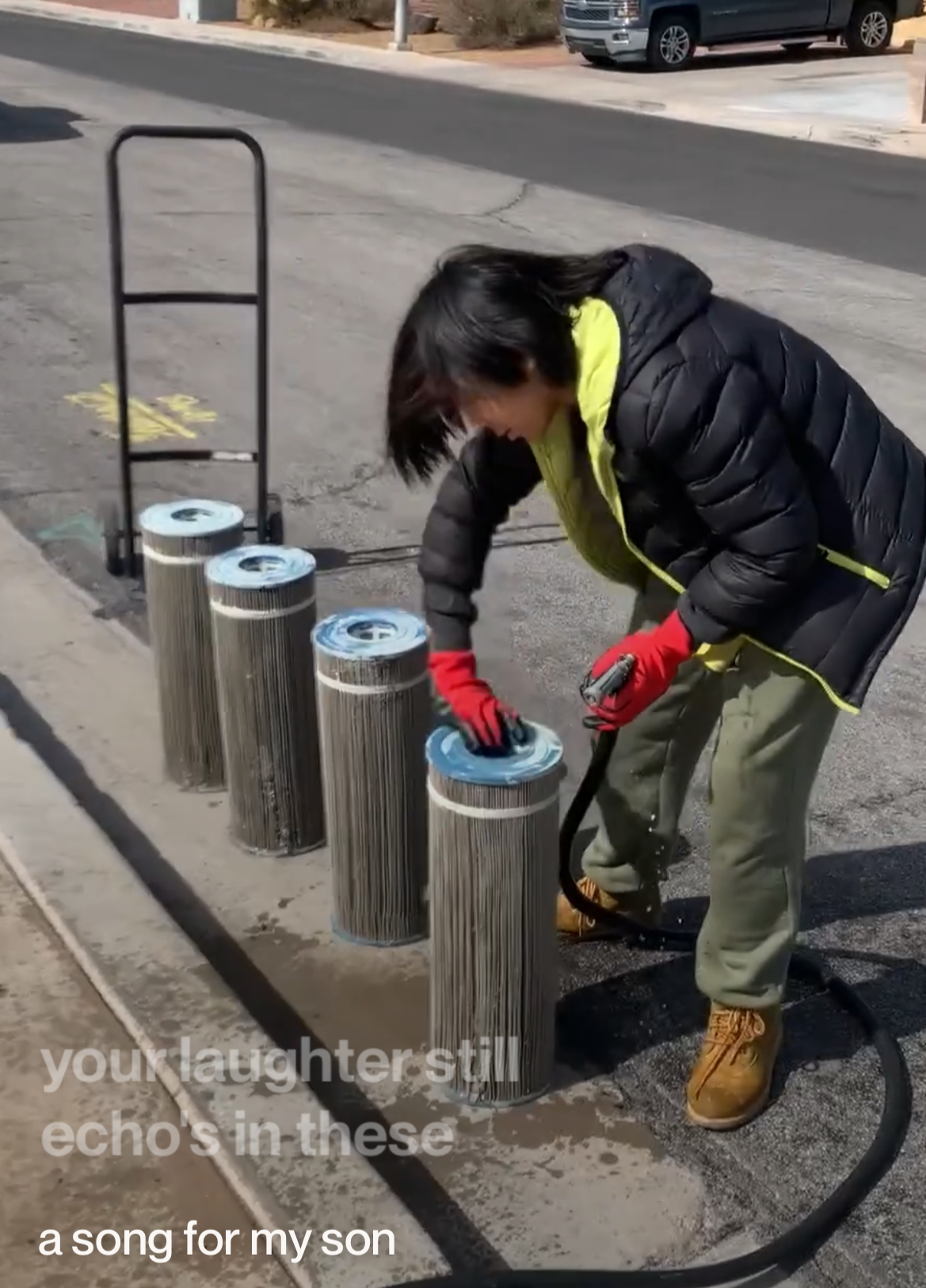 A person wearing a black jacket, red gloves, and tan boots is doing maintenance work on metal posts on a sidewalk, with a toolbox nearby and a parked vehicle in a residential area.